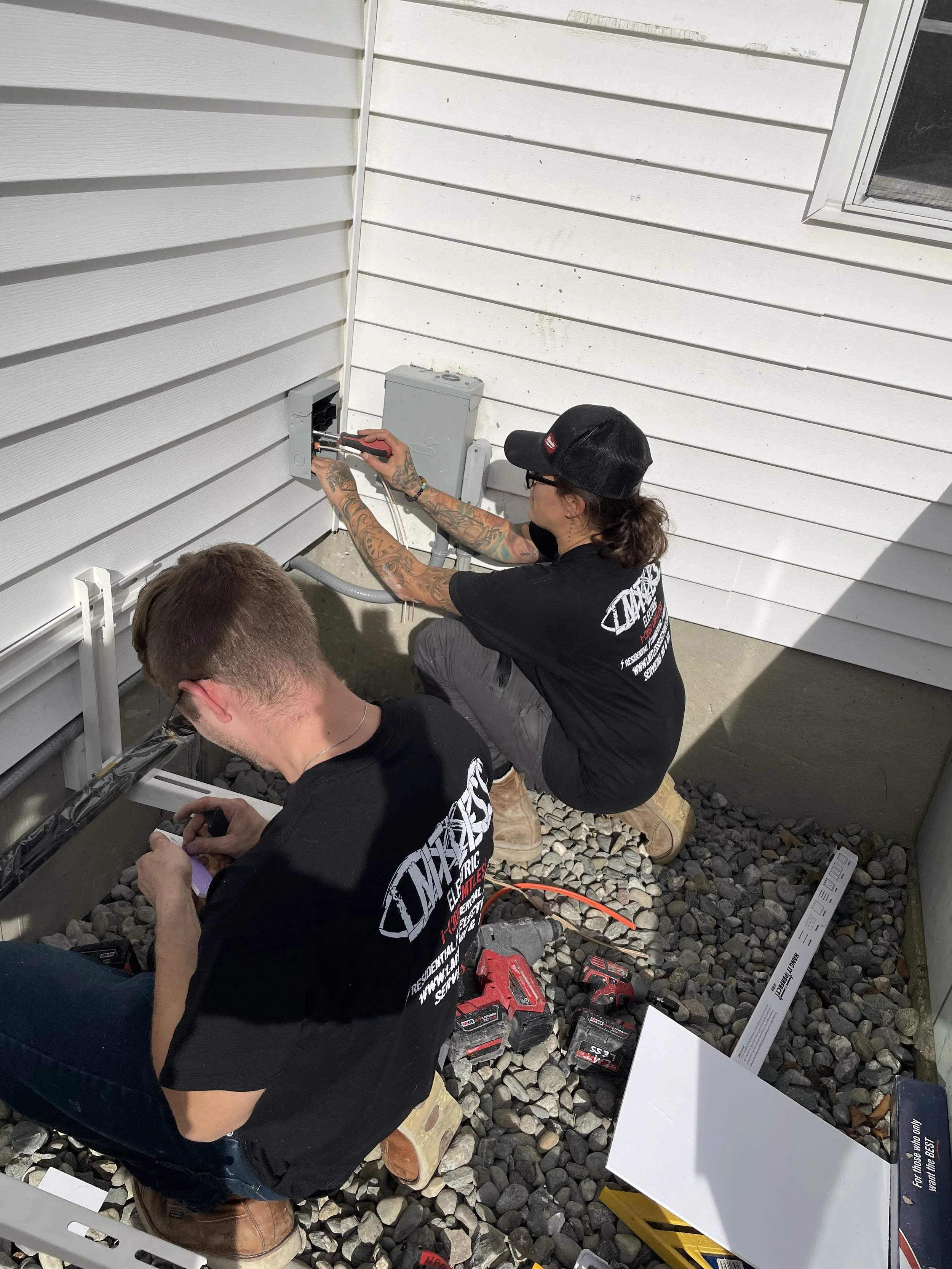 Two workers installing or repairing an electrical box on the exterior wall of a house, surrounded by tools and equipment.