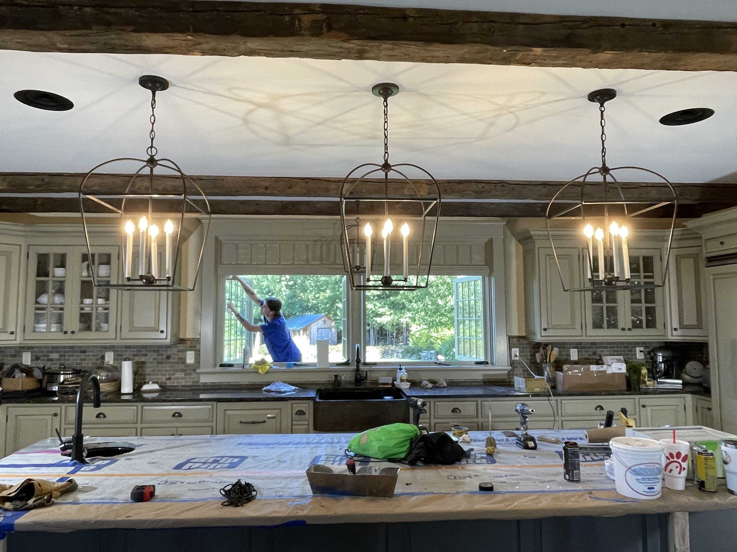 A person cleaning a window in a kitchen with cream-colored cabinets, brick backsplash, and large hanging light fixtures.