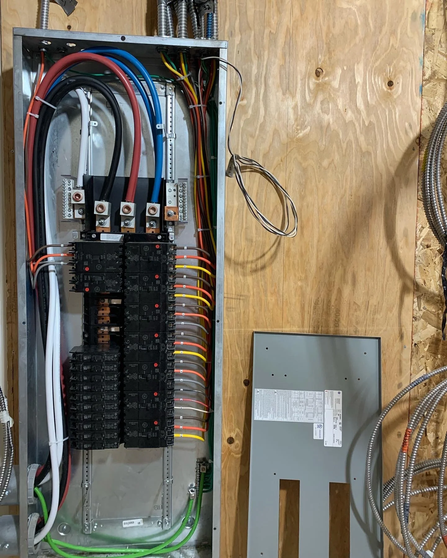 An electrical panel with circuit breakers, wires, and grounding connections mounted on a wooden wall.