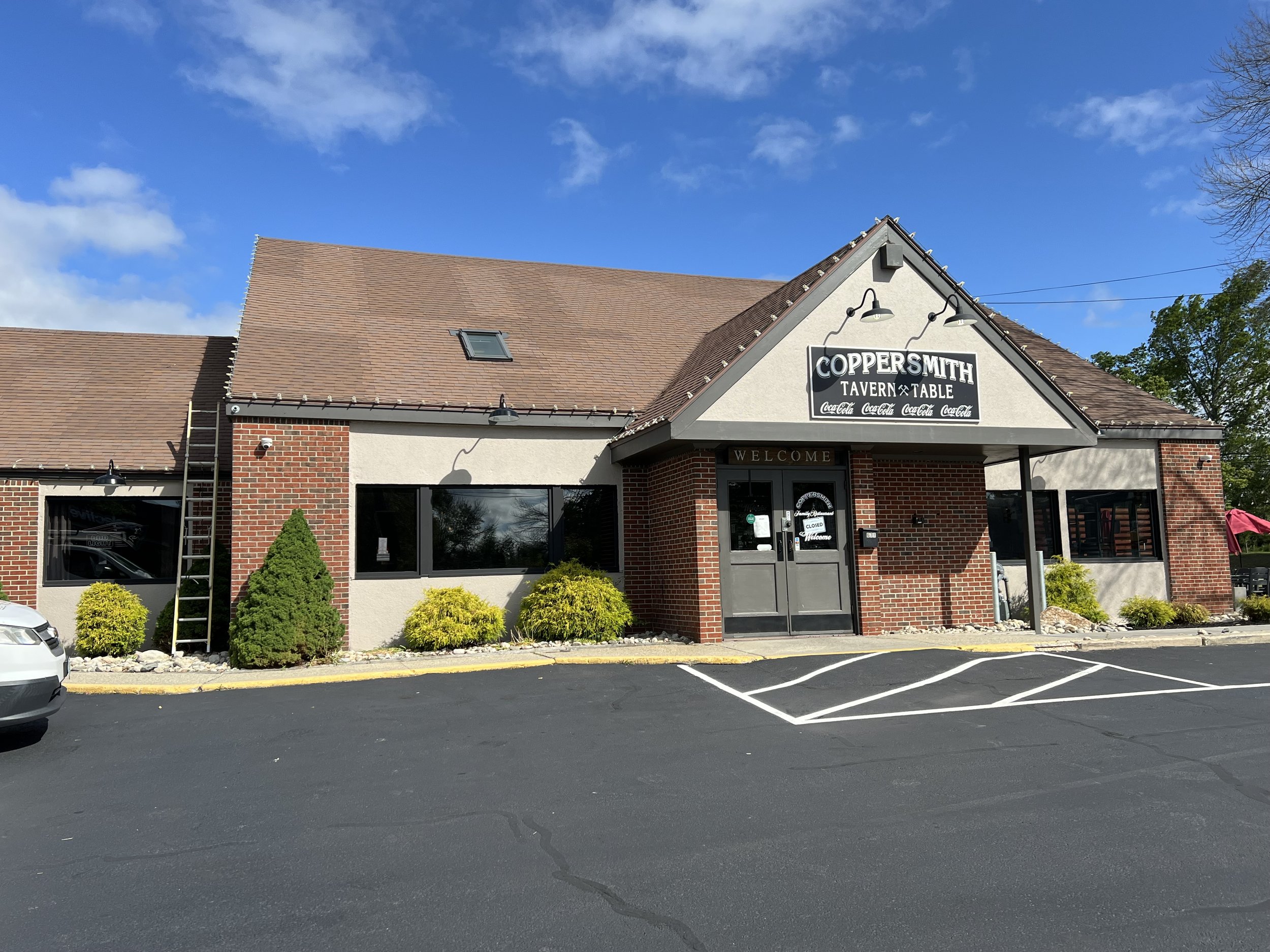 Exterior of Coppersmith Tavern & Table restaurant with a parking lot in front, landscaped bushes, a ladder on the left side, and a partly cloudy sky.