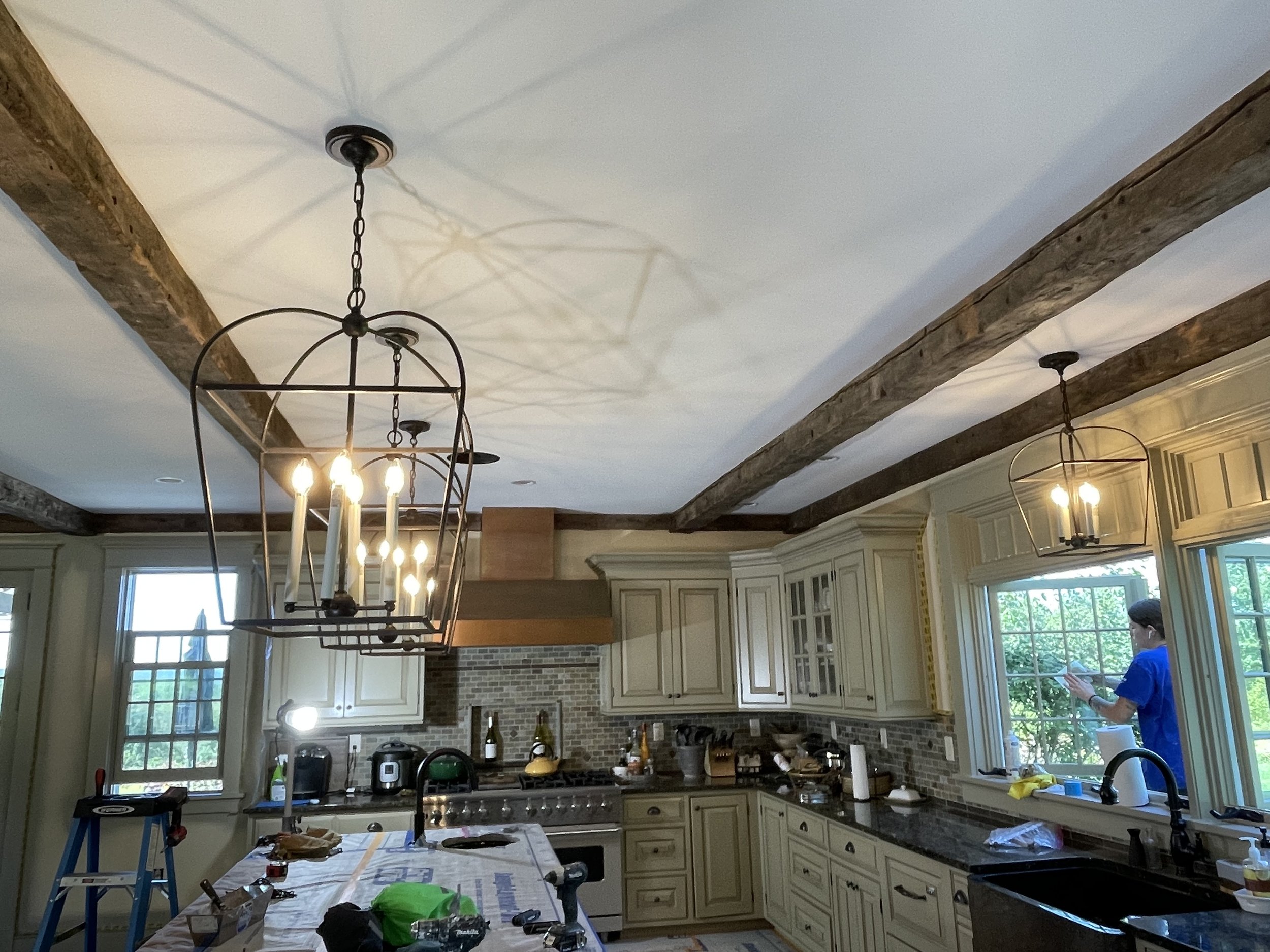 Kitchen under construction with a large chandelier hanging from a ceiling with wooden beams, kitchen cabinets, and a person working near a window.