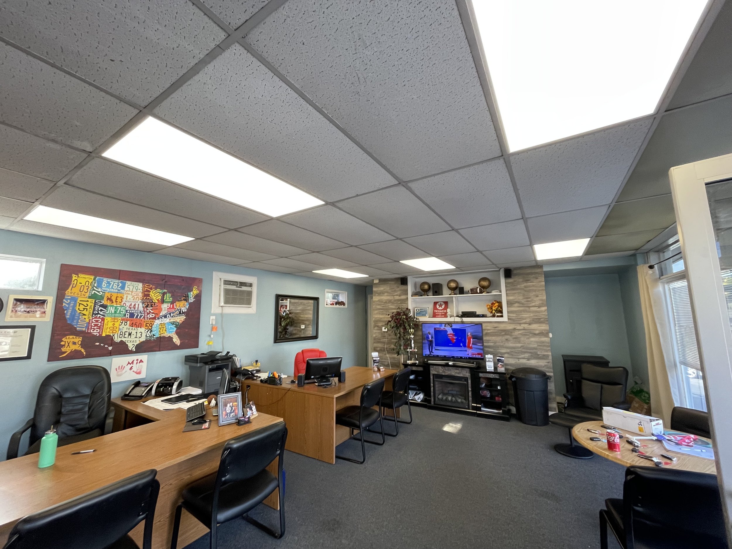 Office room with desks, chairs, a wall-mounted TV showing a weather news forecast, framed pictures, and a colorful map of the United States on the wall.