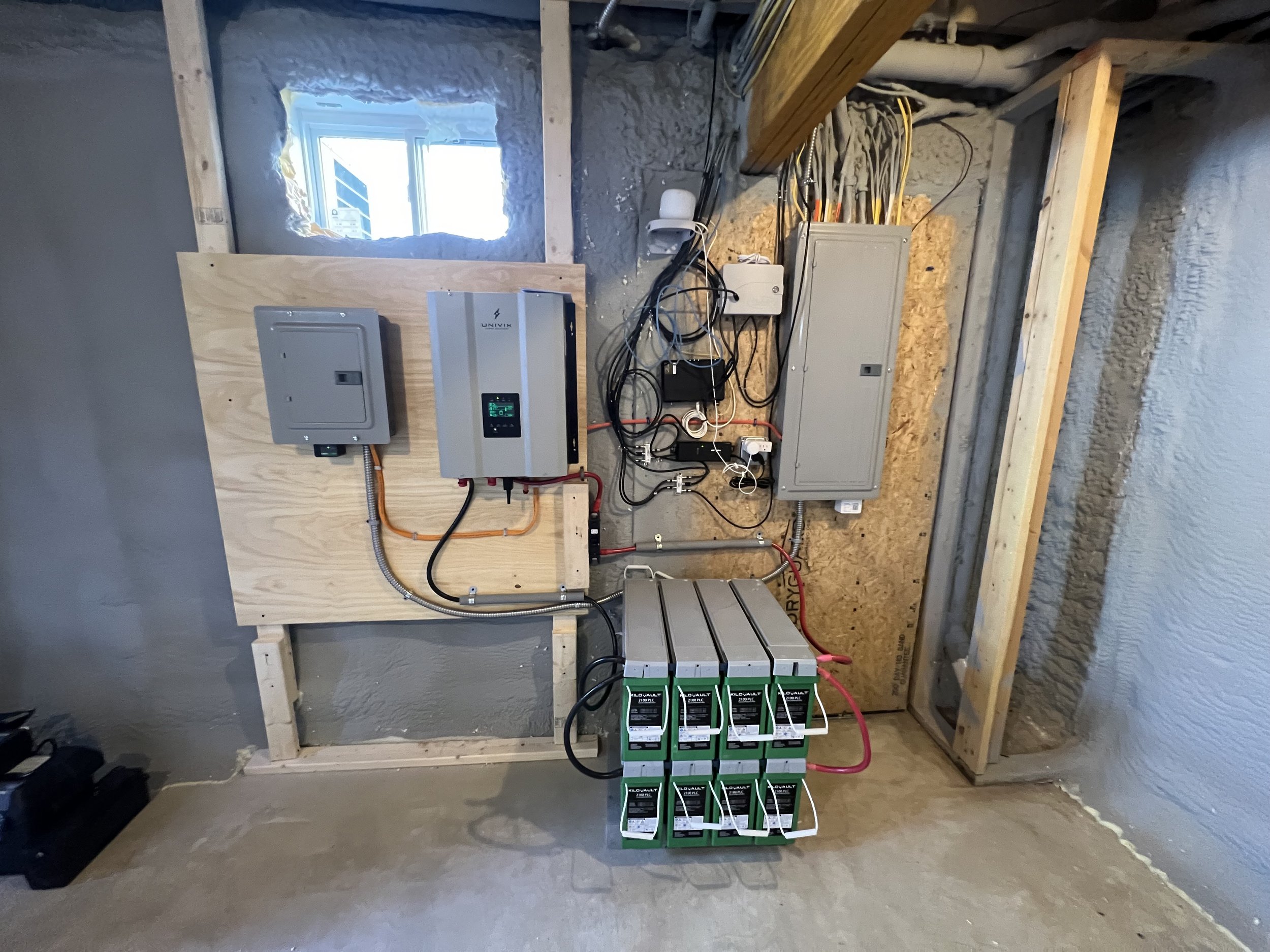 Electrical and power inverter system with batteries and control panels in an unfinished basement with a small window.