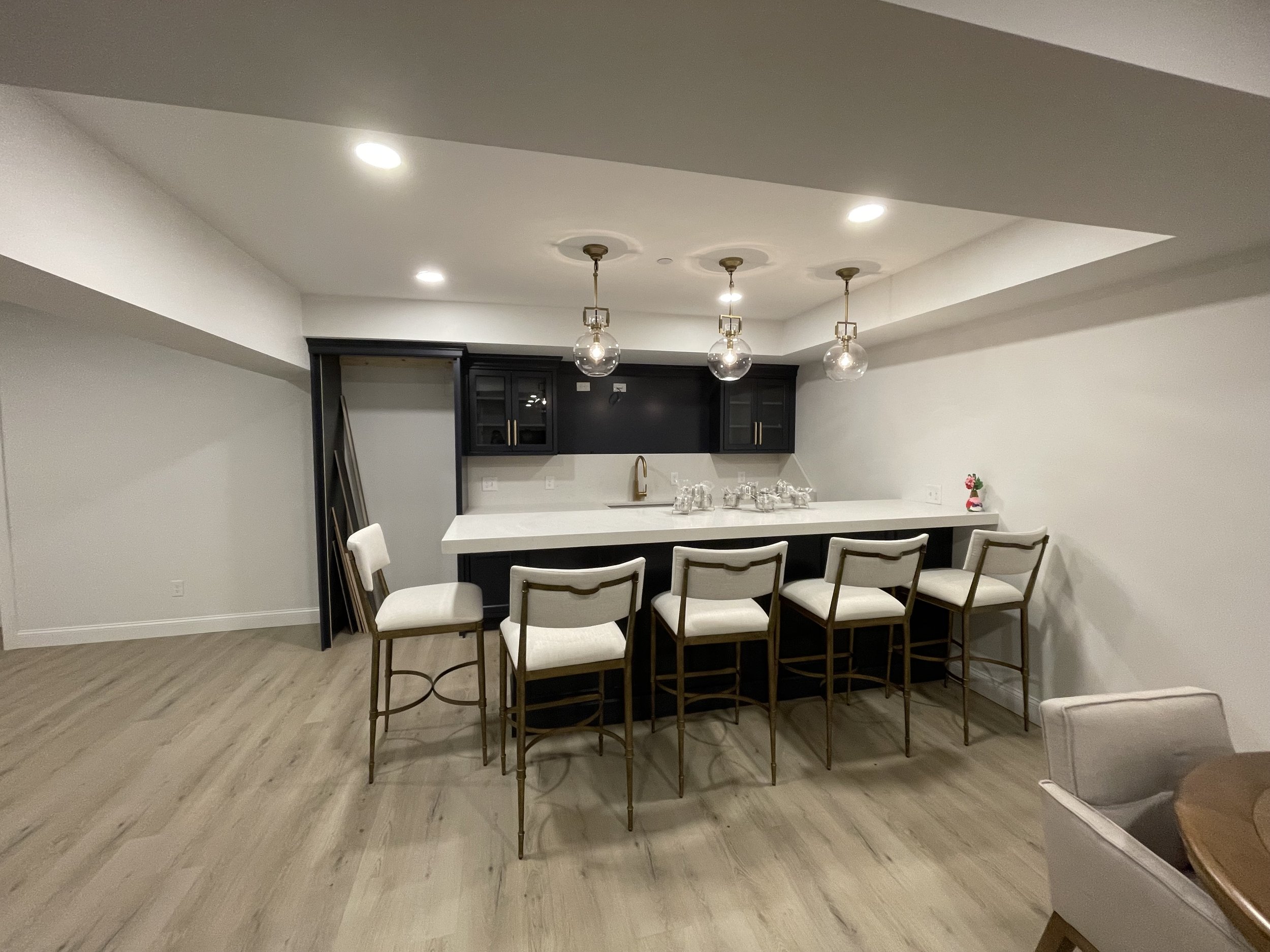 Modern kitchen area with a white countertop, black cabinets, hanging glass pendant lights, and white upholstered chairs with gold frames.