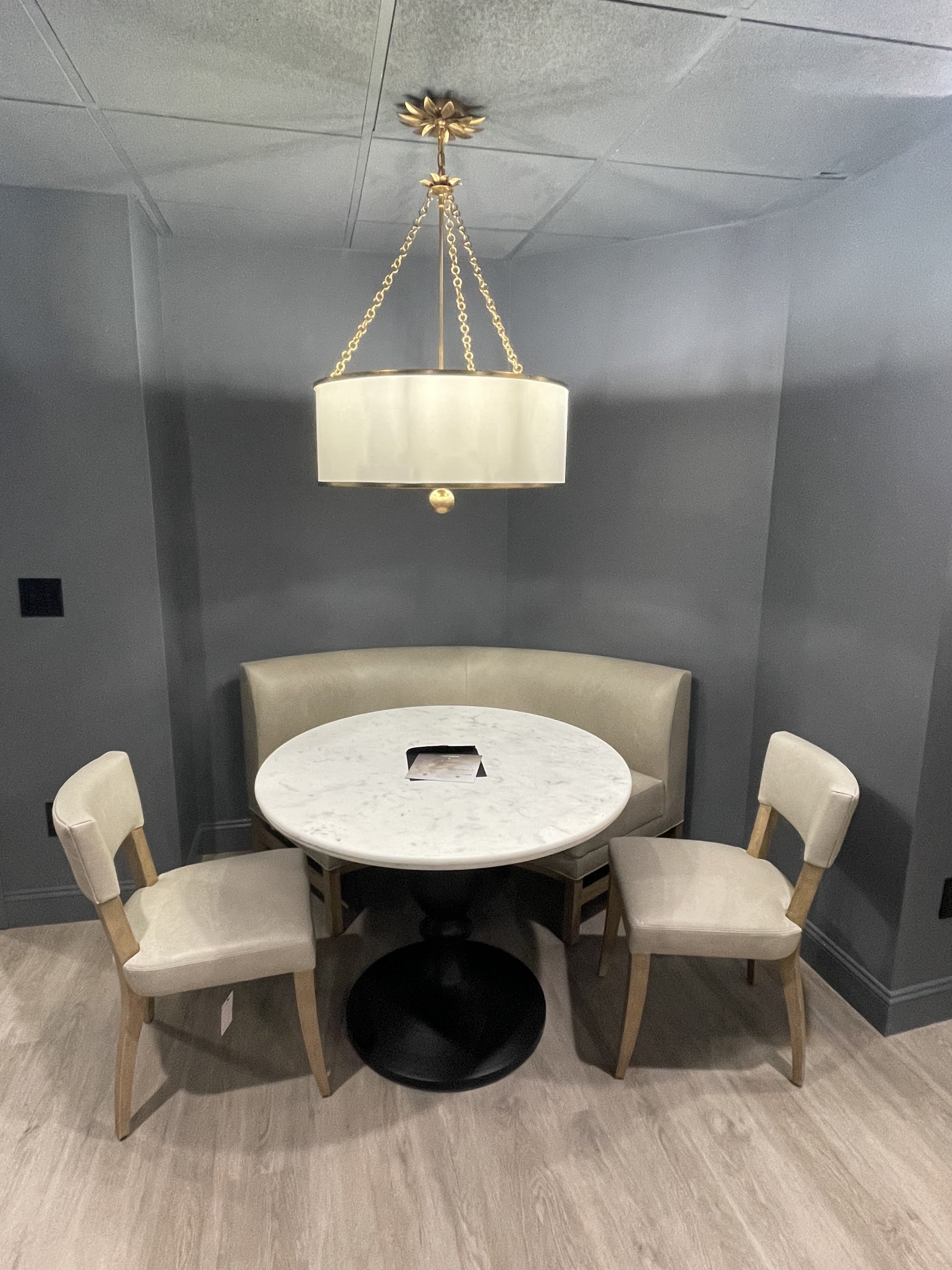 A small round table with a marble top, surrounded by three chairs with beige cushions, and a curved beige banquette seating against a dark gray wall, illuminated by a large pendant light with a cream-colored shade hanging from the ceiling.