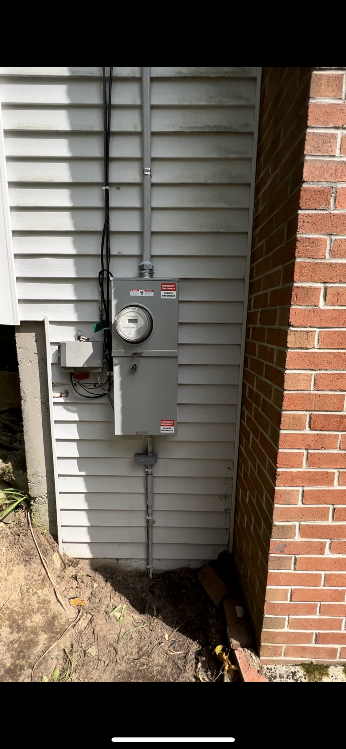 Exterior view of an electric meter box attached to the house wall, with electrical wires and a service disconnect label, next to a brick wall and dirt ground.