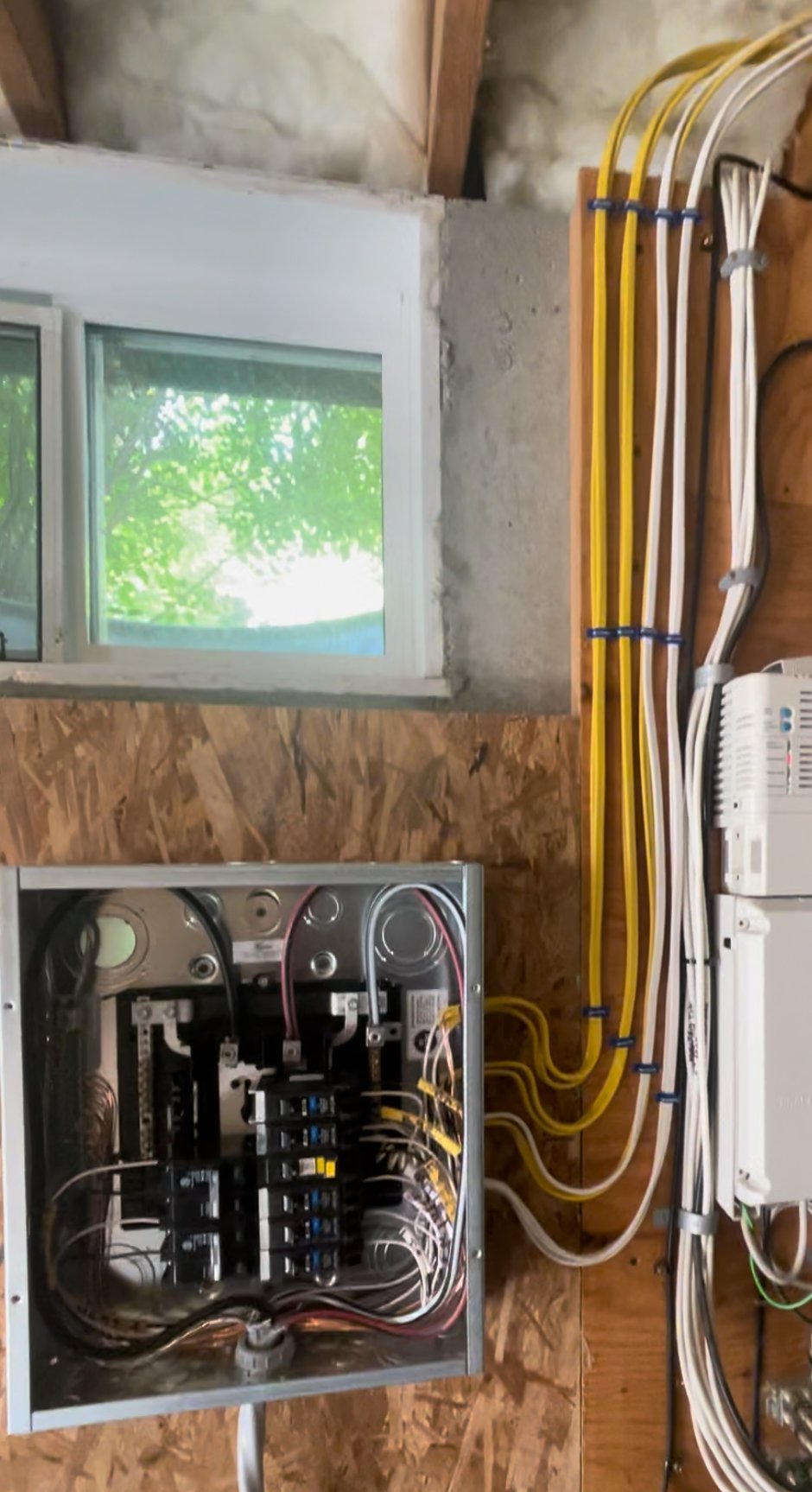 Electrical wiring and breaker box mounted inside an unfinished wooden wall near a window with green trees outside.
