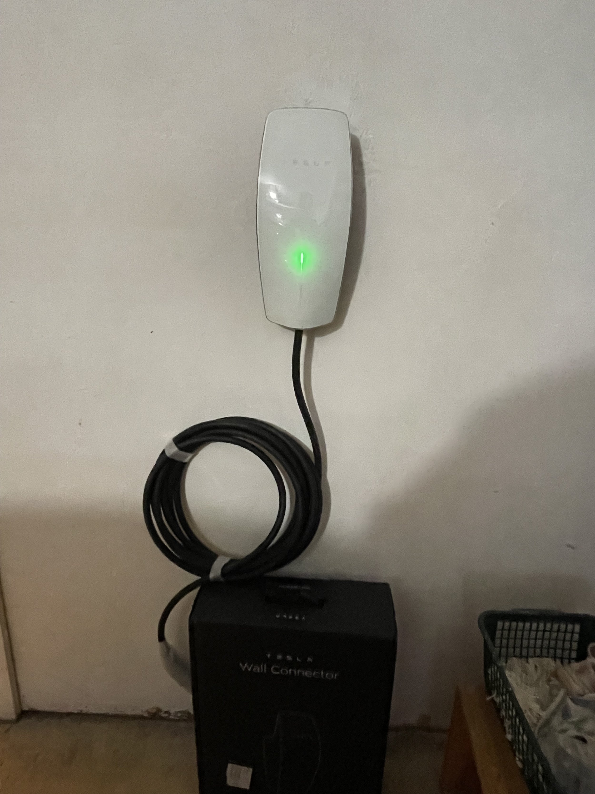 Electric vehicle wall charger mounted on a wall, connected to a black wall outlet box with a coiled charging cable.