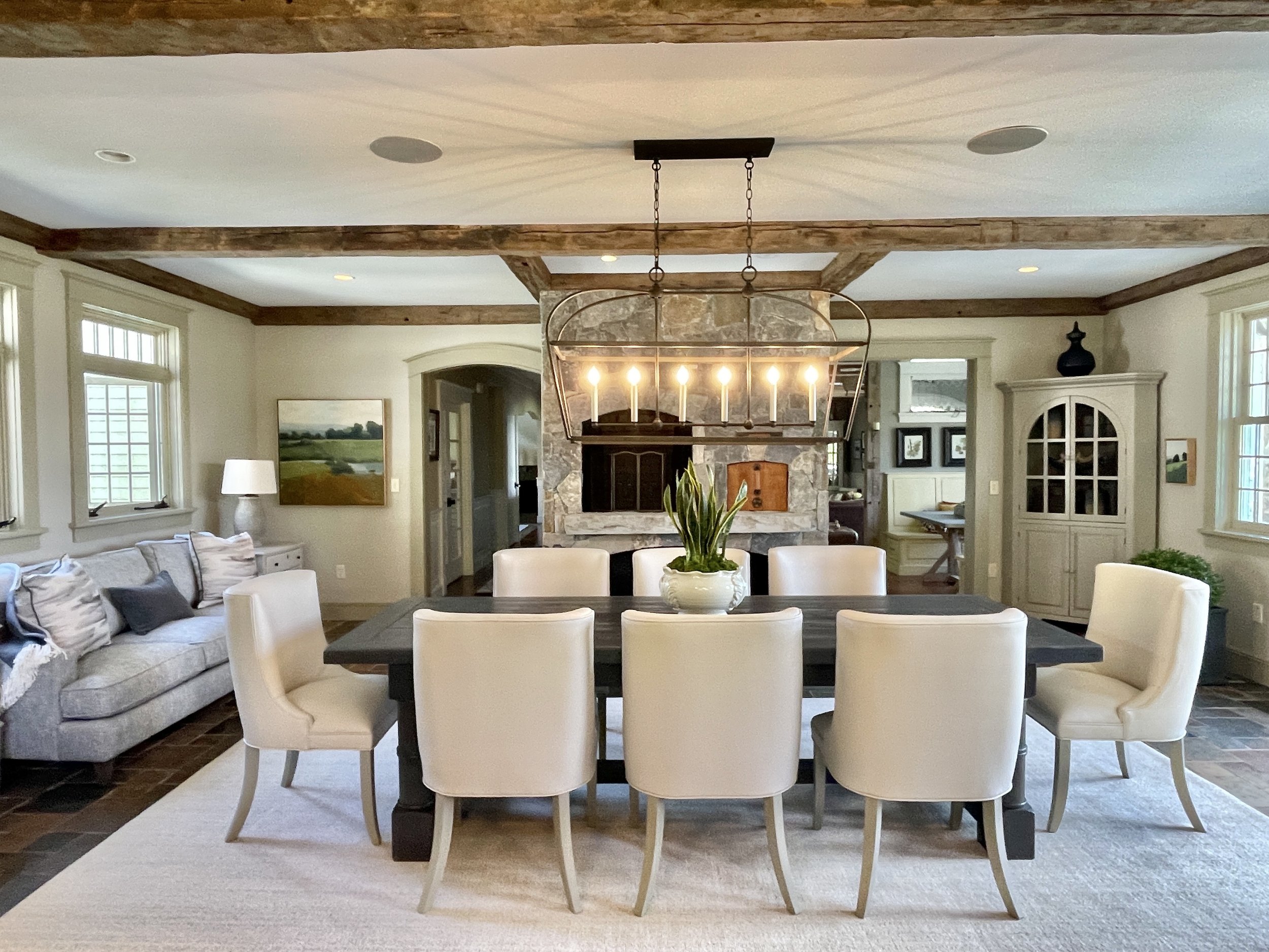 Dining room with a large black table surrounded by eight cream-colored chairs, a stone fireplace, a hanging chandelier with candles, a gray sofa with pillows, and large windows letting in natural light.