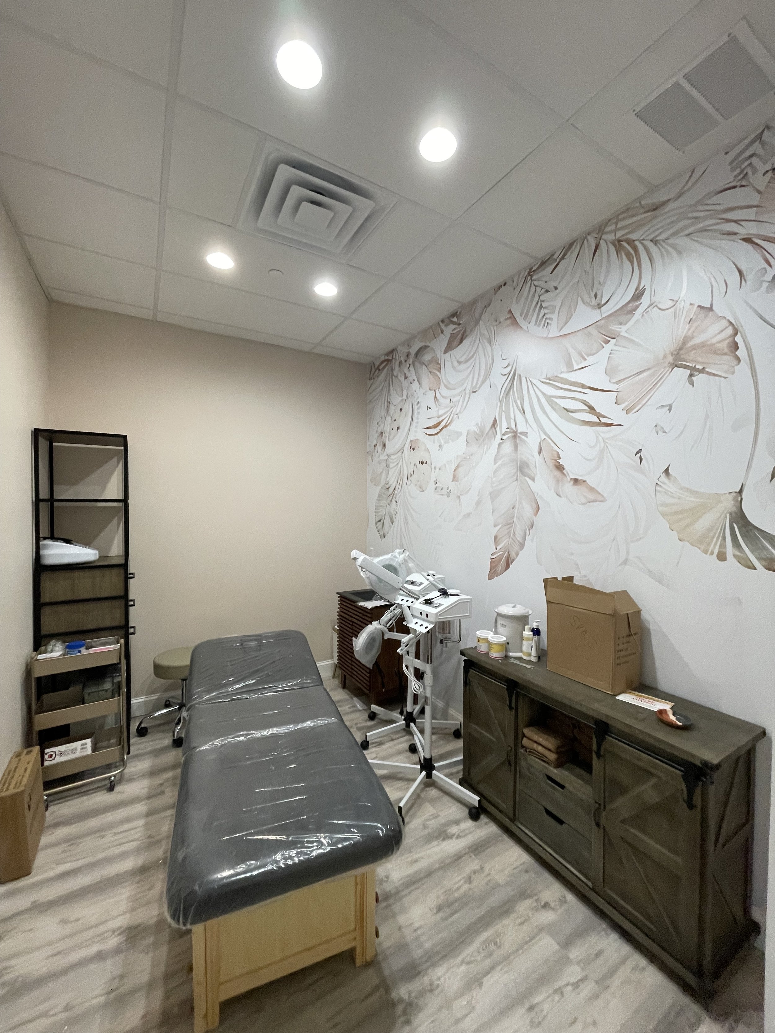 Medical treatment room with a massage or examination table covered in plastic, a rolling stool, a cabinet on the right with supplies, a shelf on the left, and a wall with a large floral mural.