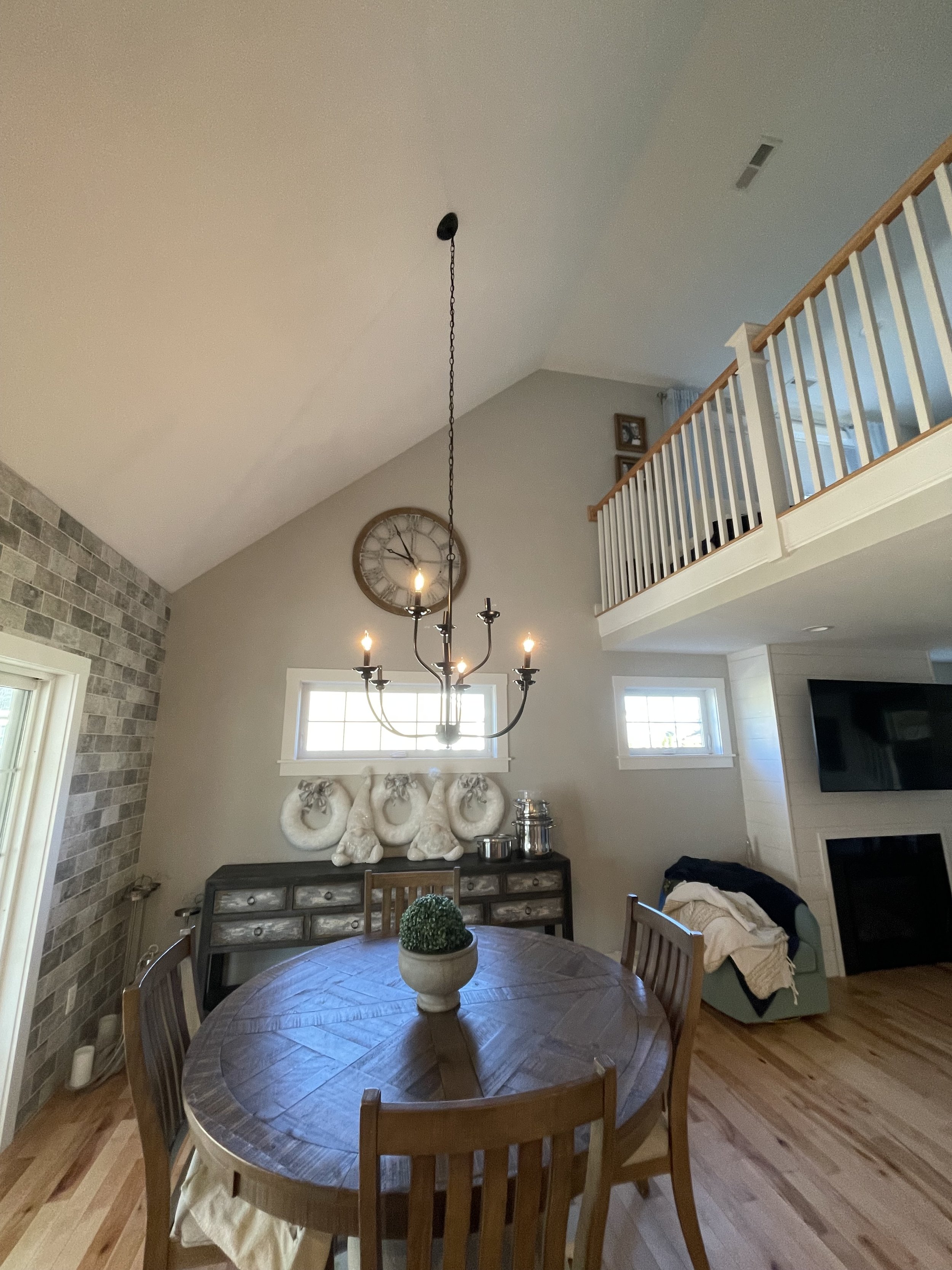 A dining room with a round wooden table, a black buffet, wall decorations, and a chandelier hanging from a high ceiling, with a loft area above.