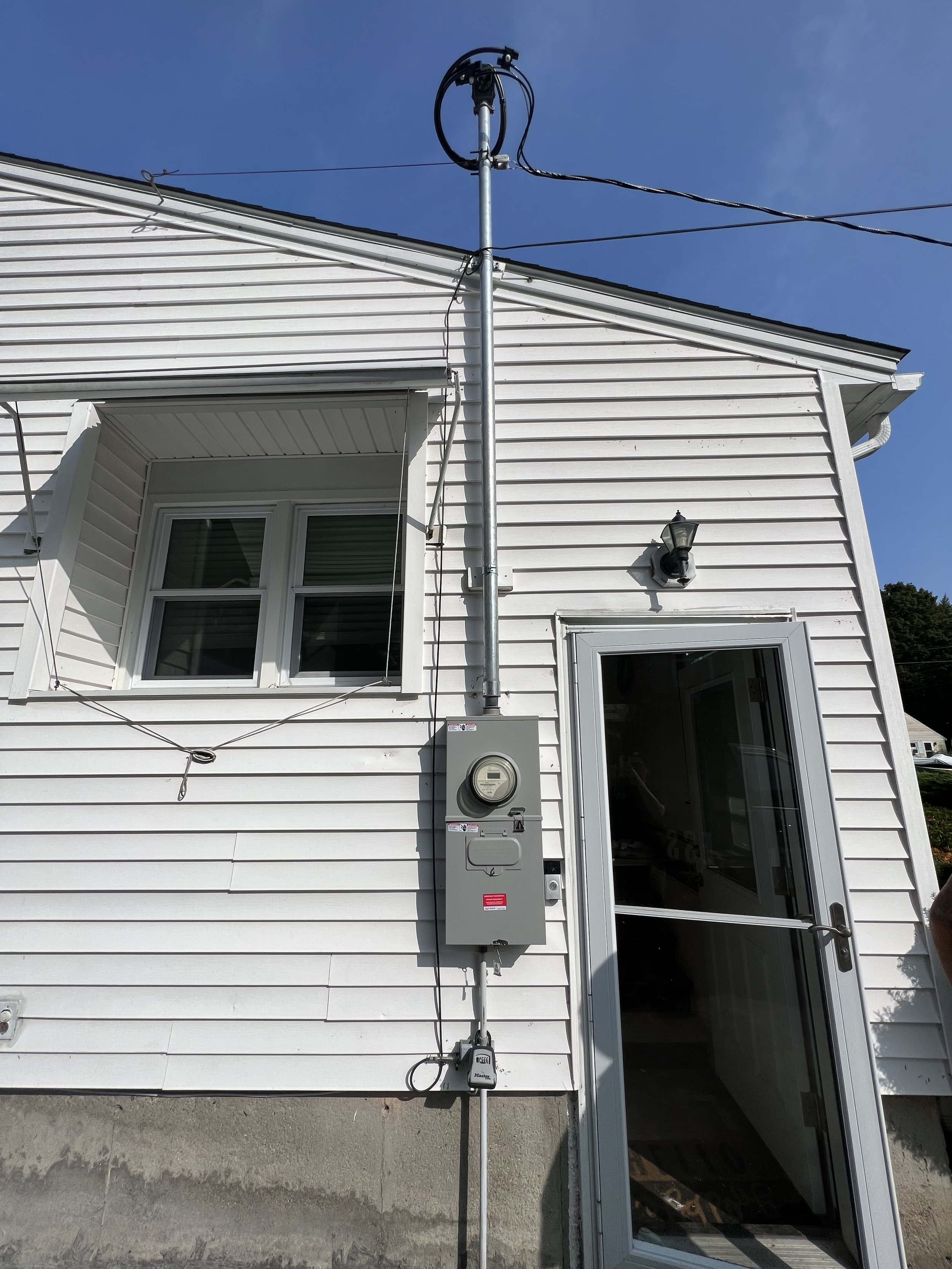 Exterior of a white house with a window, door, and an electric meter box mounted on the wall. An outdoor lamp is next to the door, and electrical wires run from the pole above.