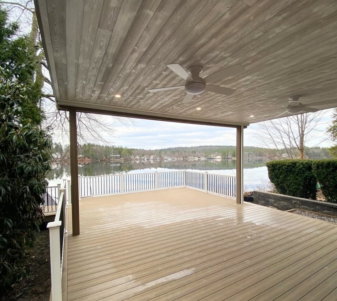 Covered wooden deck overlooking a lake with a white railing, surrounded by trees and shrubs.