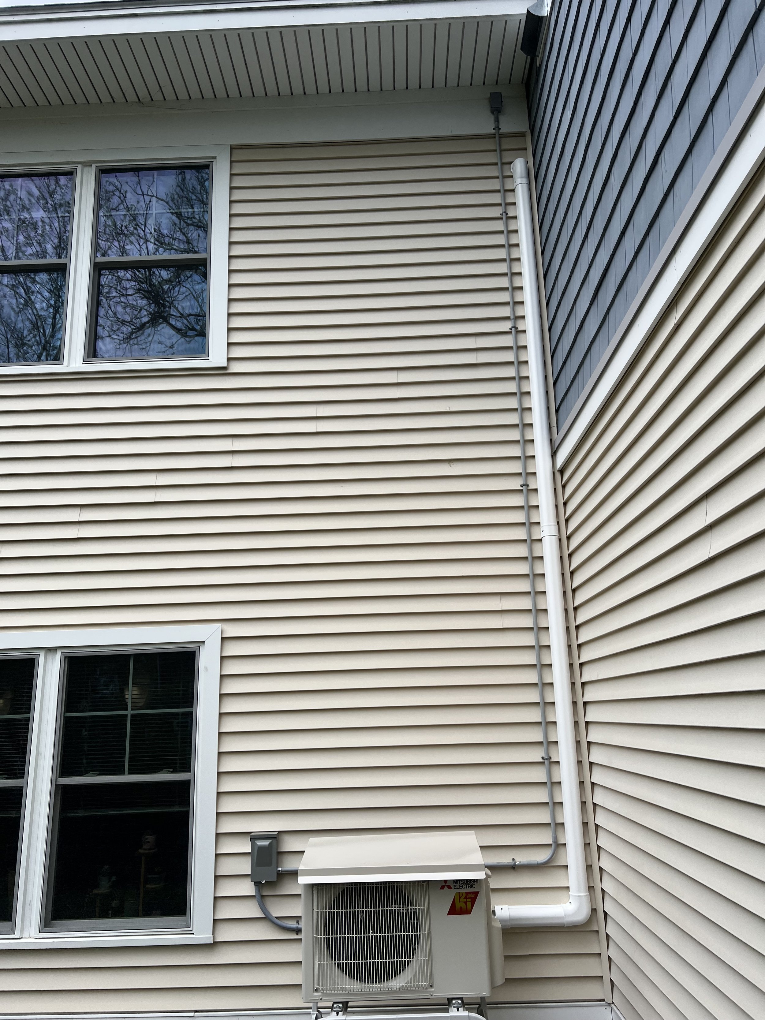 Exterior view of a house with beige and blue siding, two windows, and an air conditioning unit mounted on the wall with electrical connections and piping.