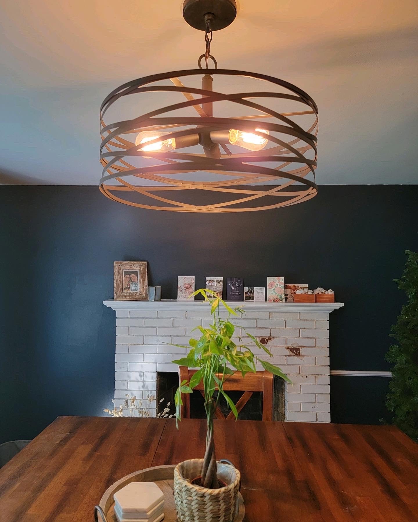 Modern chandelier hanging above a wooden dining table with a potted plant and framed photos on a white brick fireplace in the background.
