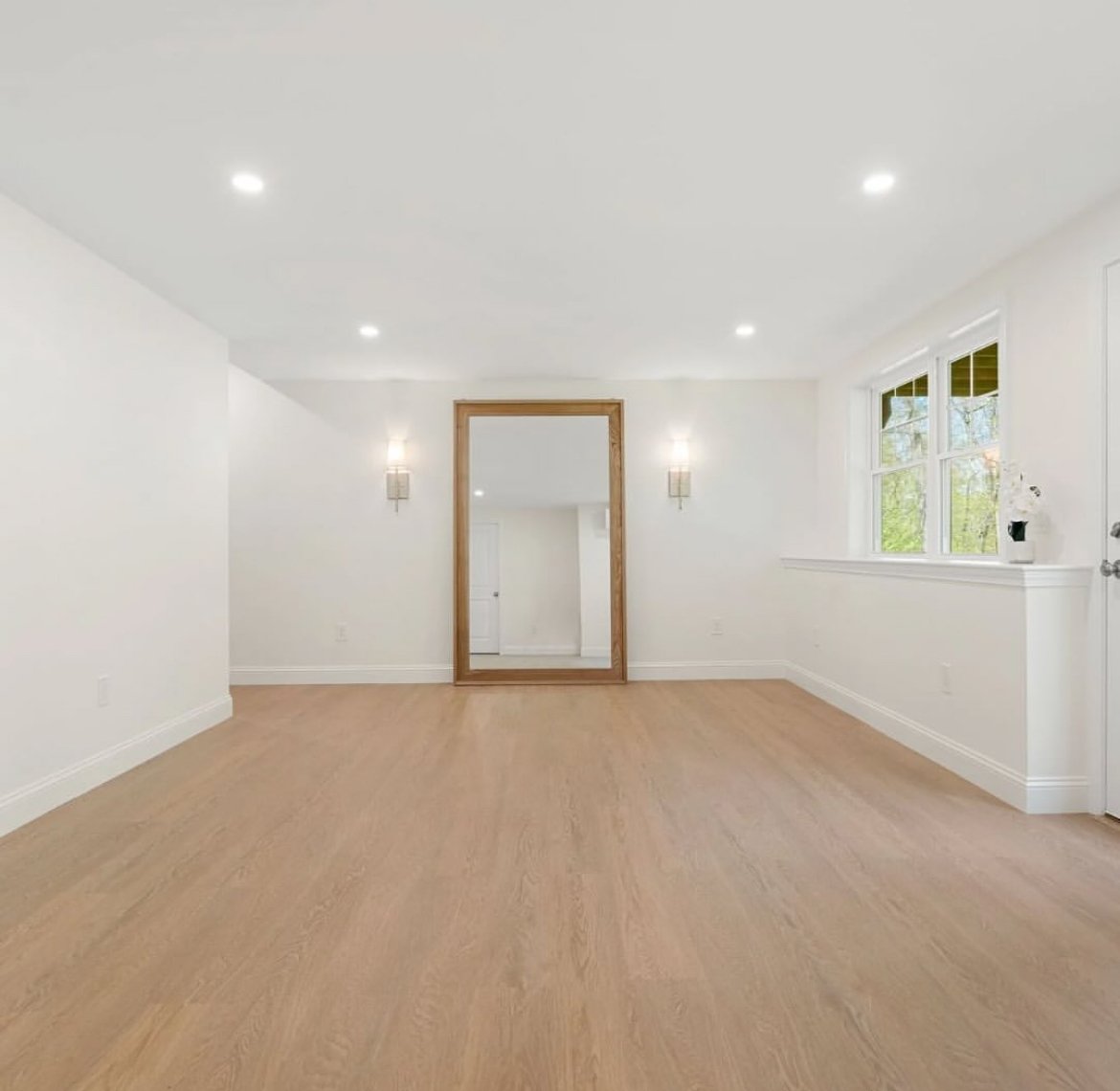 Empty room with white walls, hardwood floors, a large mirror with a wooden frame, and a window with a flowerpot on the windowsill.