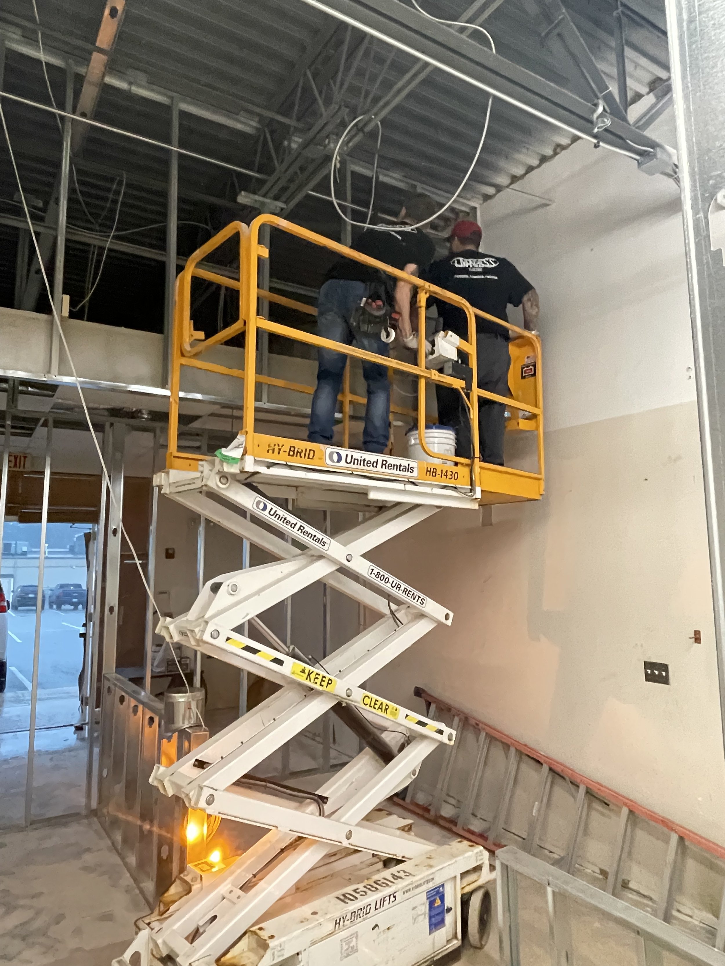 Two workers are on an orange and white scissor lift installing or repairing ceiling fixtures in a commercial space.