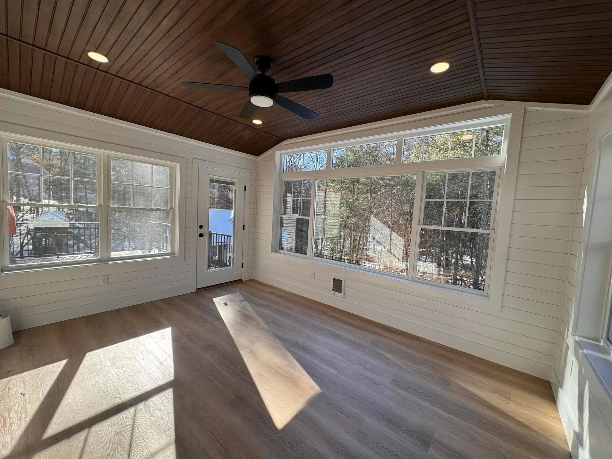 Sunlit enclosed porch or sunroom with large windows, wood floors, white wall paneling, a black ceiling fan, and a wooden ceiling.
