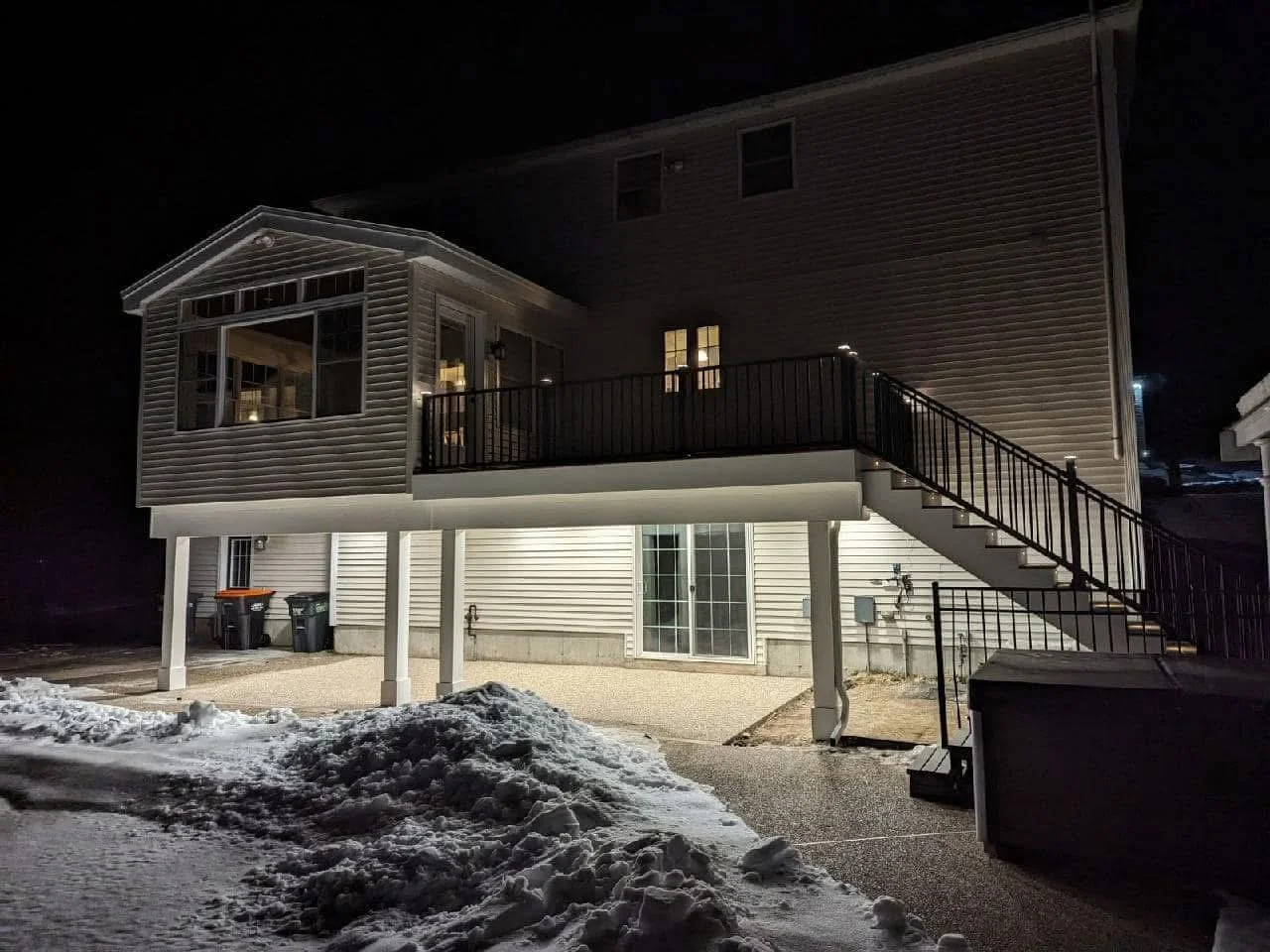 Nighttime view of a two-story house with a raised deck, stairs, and lit interior, snow on the ground, and garbage bins outside.