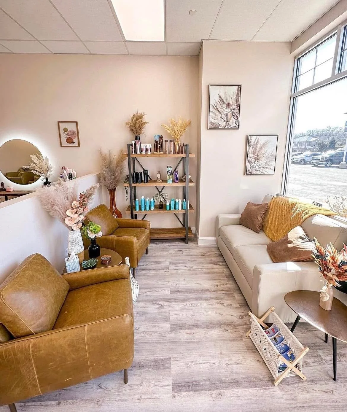 A cozy waiting area with two brown leather chairs, a white sofa, a small side table with decorative flowers, a black shelf with skincare products, framed floral artwork, and a large window letting in natural light.