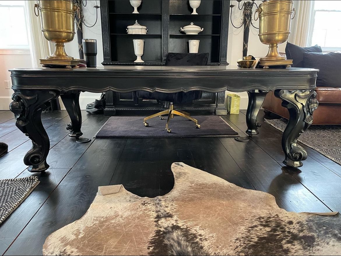 Black wooden desk with ornate curved legs in a living room with dark hardwood floors, a cowhide rug, and a black bookshelf behind the desk with white decorative vases.