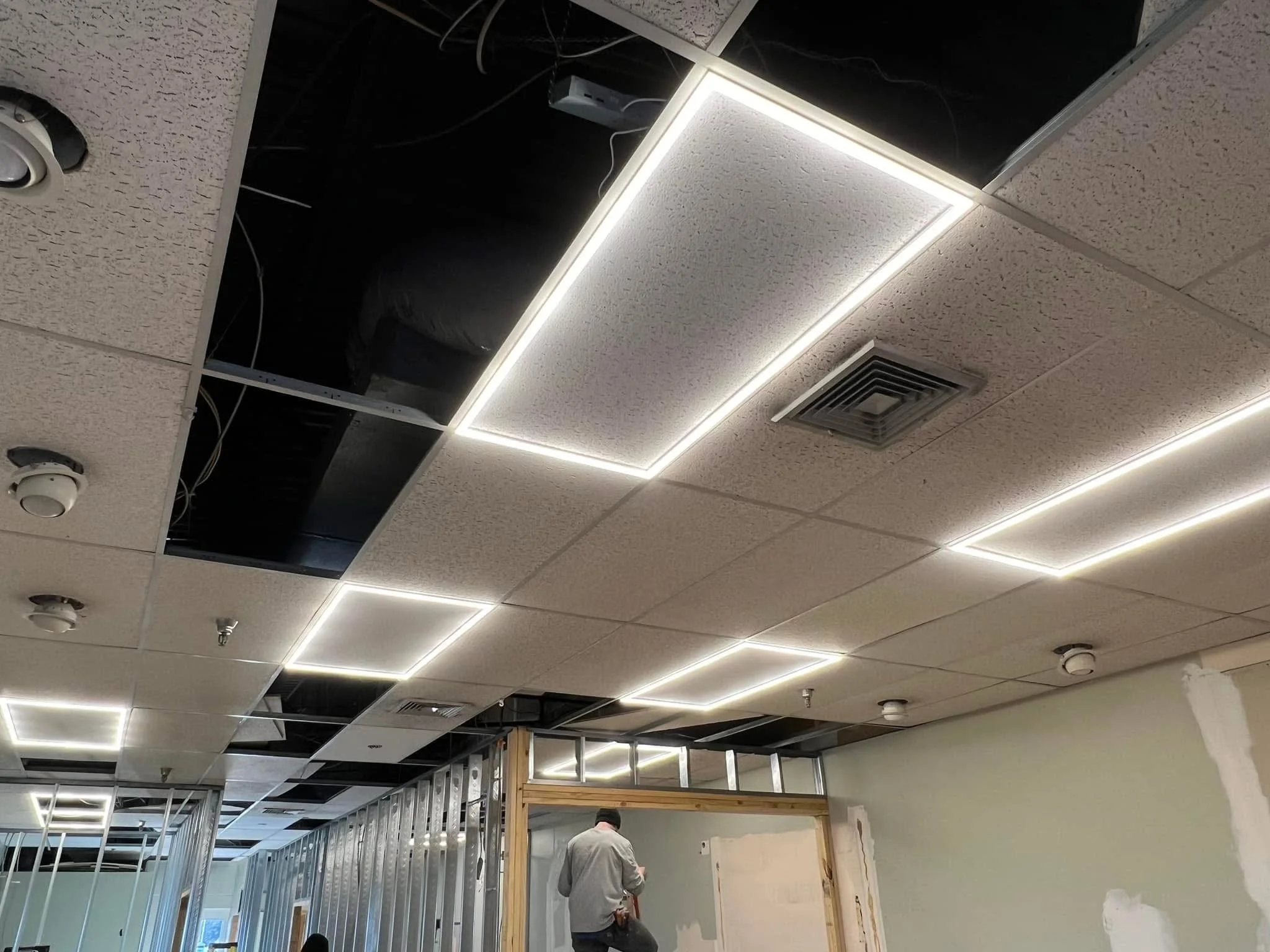 Construction worker in a room with a partially finished ceiling, some panels missing, and installed LED ceiling lights.