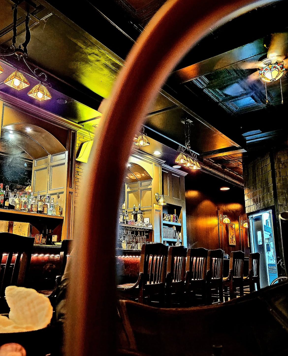 Interior of a dimly lit bar with wooden chairs and a stone accent wall, bottles of liquor behind the bar, and warm overhead lighting.