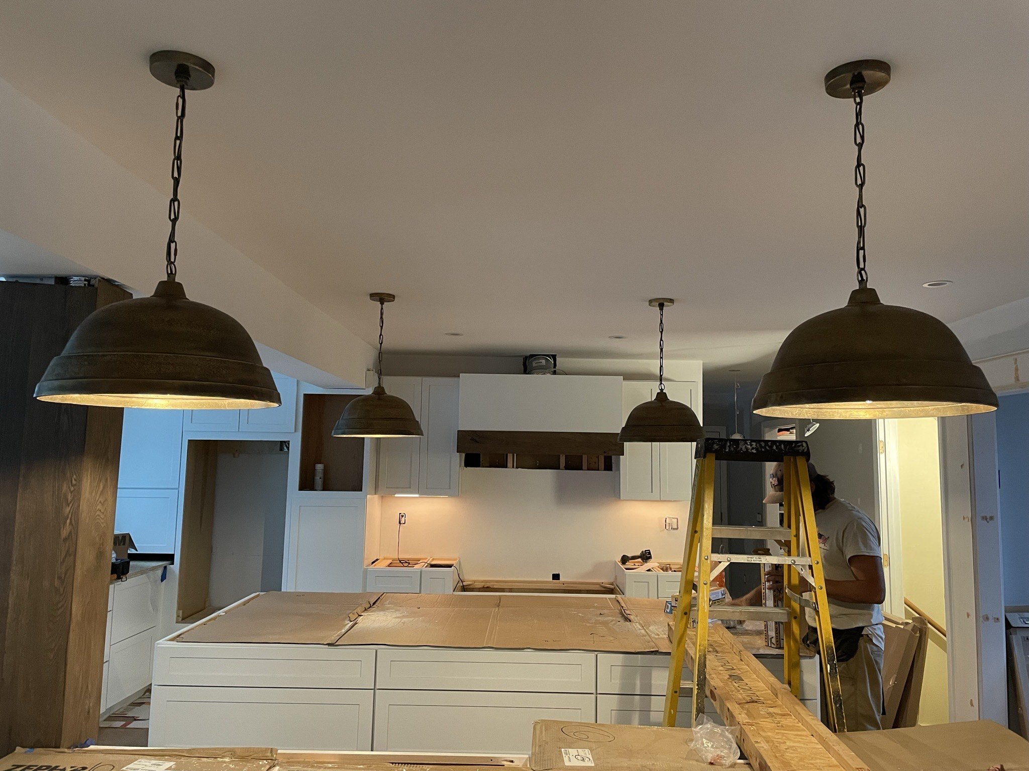 Kitchen under renovation with white cabinets, a yellow ladder, and workers installing light fixtures.