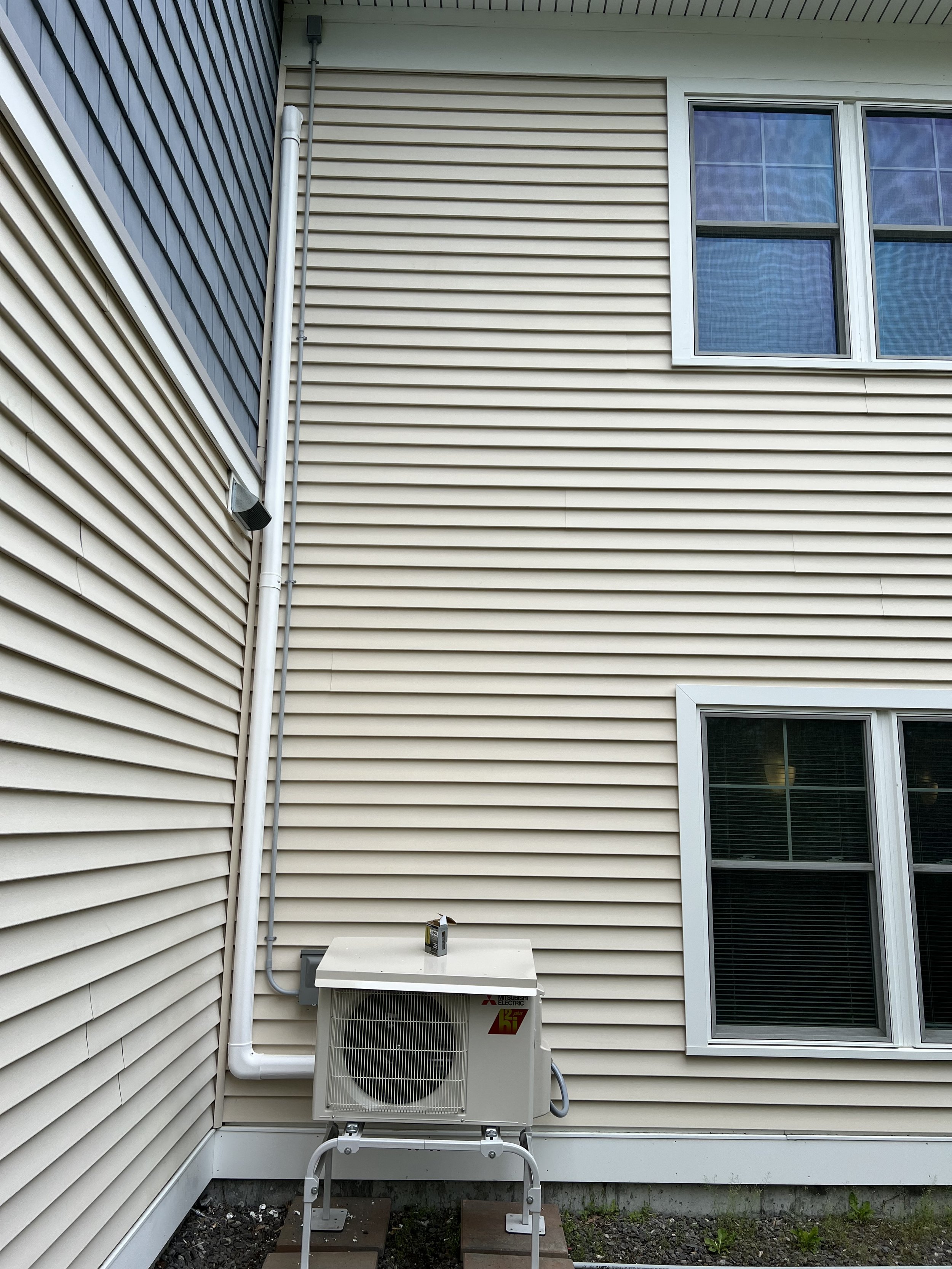 Exterior view of a house with beige vinyl siding, showing a window, an air conditioning unit, and visible plumbing pipes.