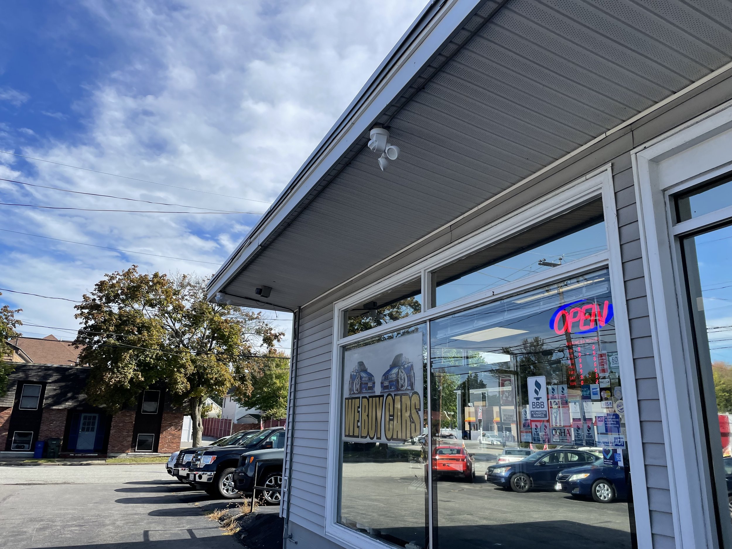 Car dealership with a large window sign reading "WE BUY CARS" and a neon "OPEN" sign inside. Several parked cars are visible outside, along with a building, trees, and a partly cloudy sky.