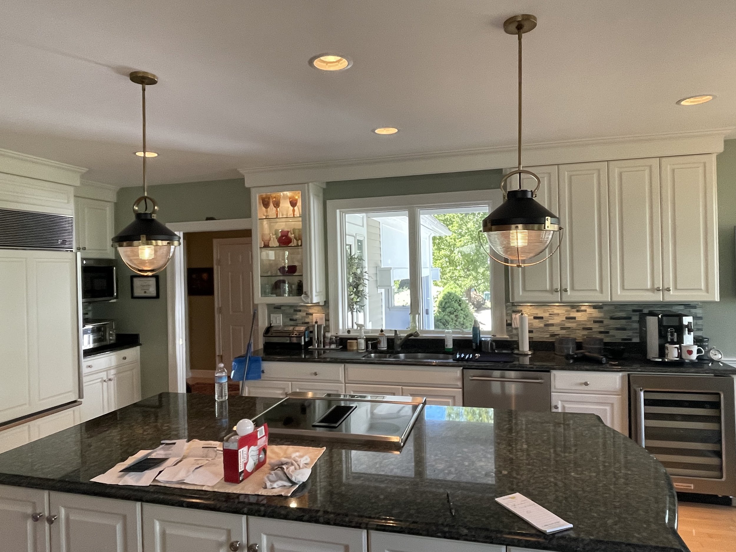 Modern kitchen with white cabinets, black granite countertops, pendant lights, a large window, and various appliances including a microwave, coffee maker, and wine cooler. The island has a water bottle, paper towels, and some packaging on it.