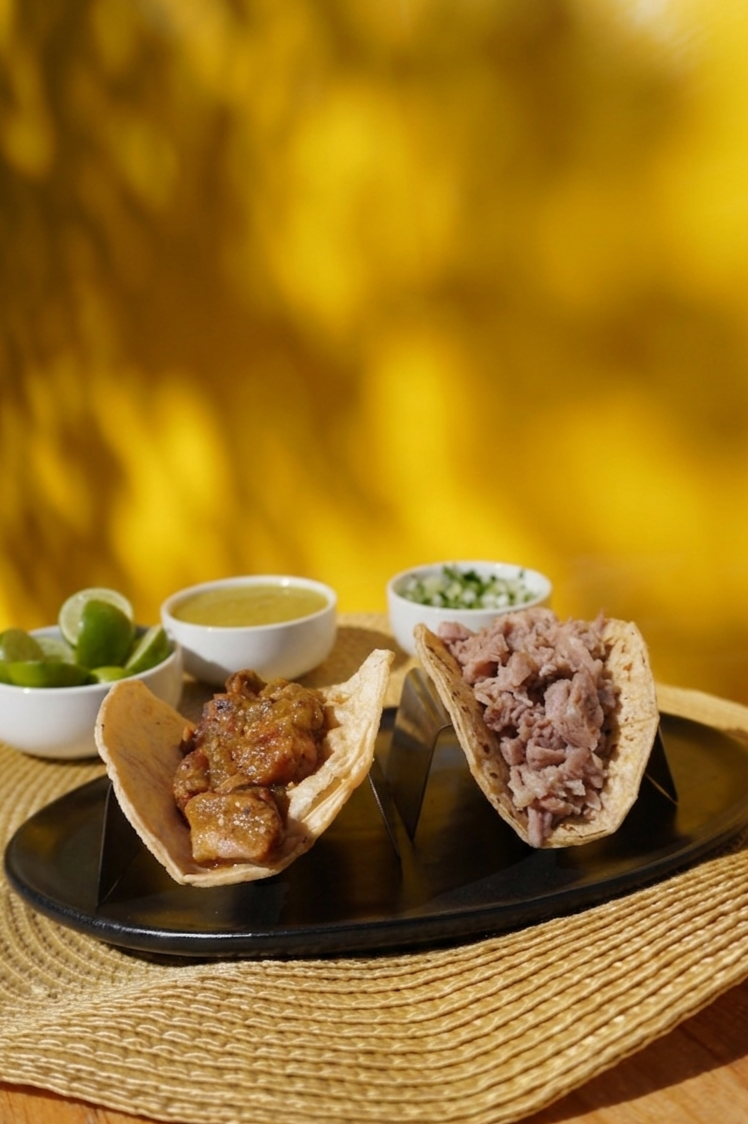 Two tacos with shredded meat on corn tortillas, served with lime, chopped onions, and green sauce on the side, on a black plate with a woven placemat.