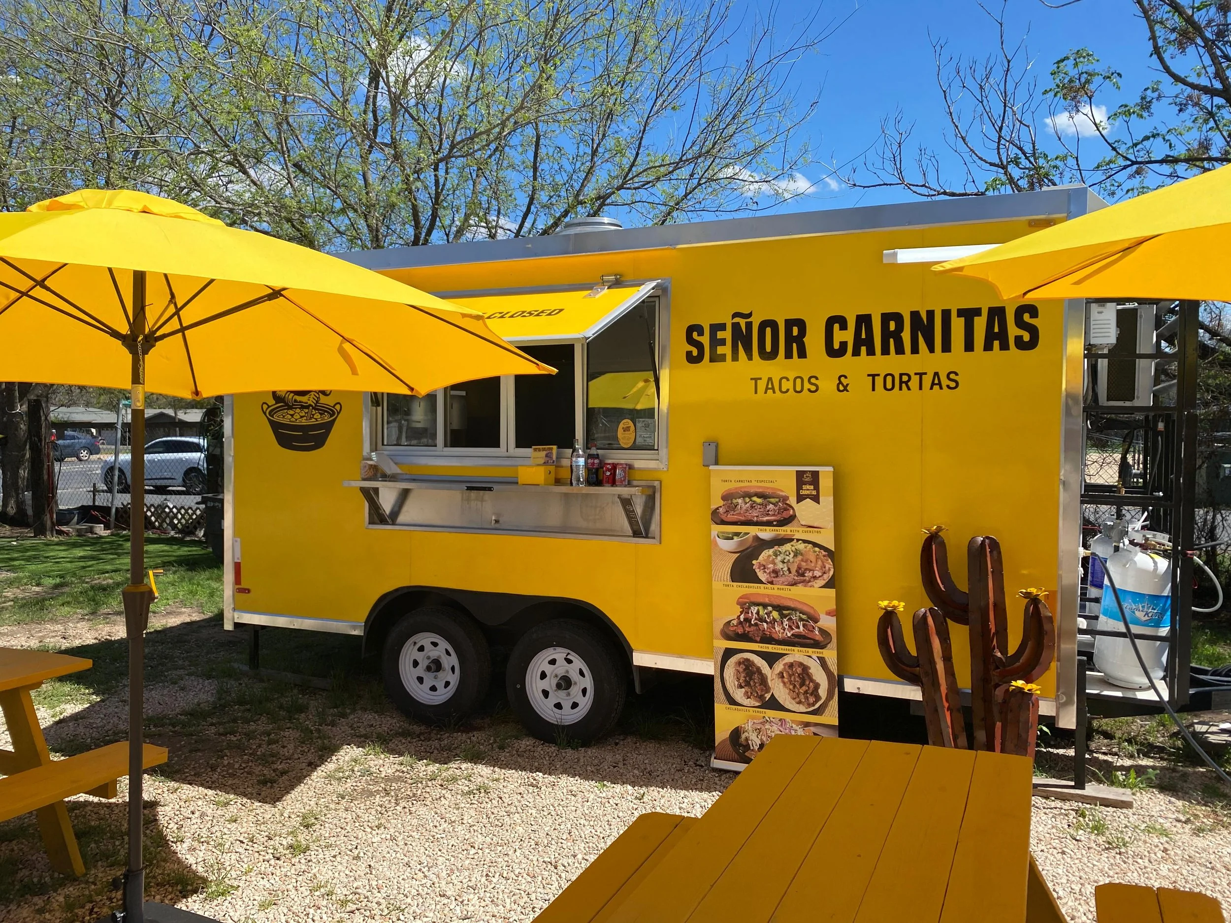One yellow umbrella two yellow picnic tables and a yellow food trailer with a small brown cactus with yellow flowers and a banner displaying foos like tacos and tortas, in the background you can see a blue sky and a tree.