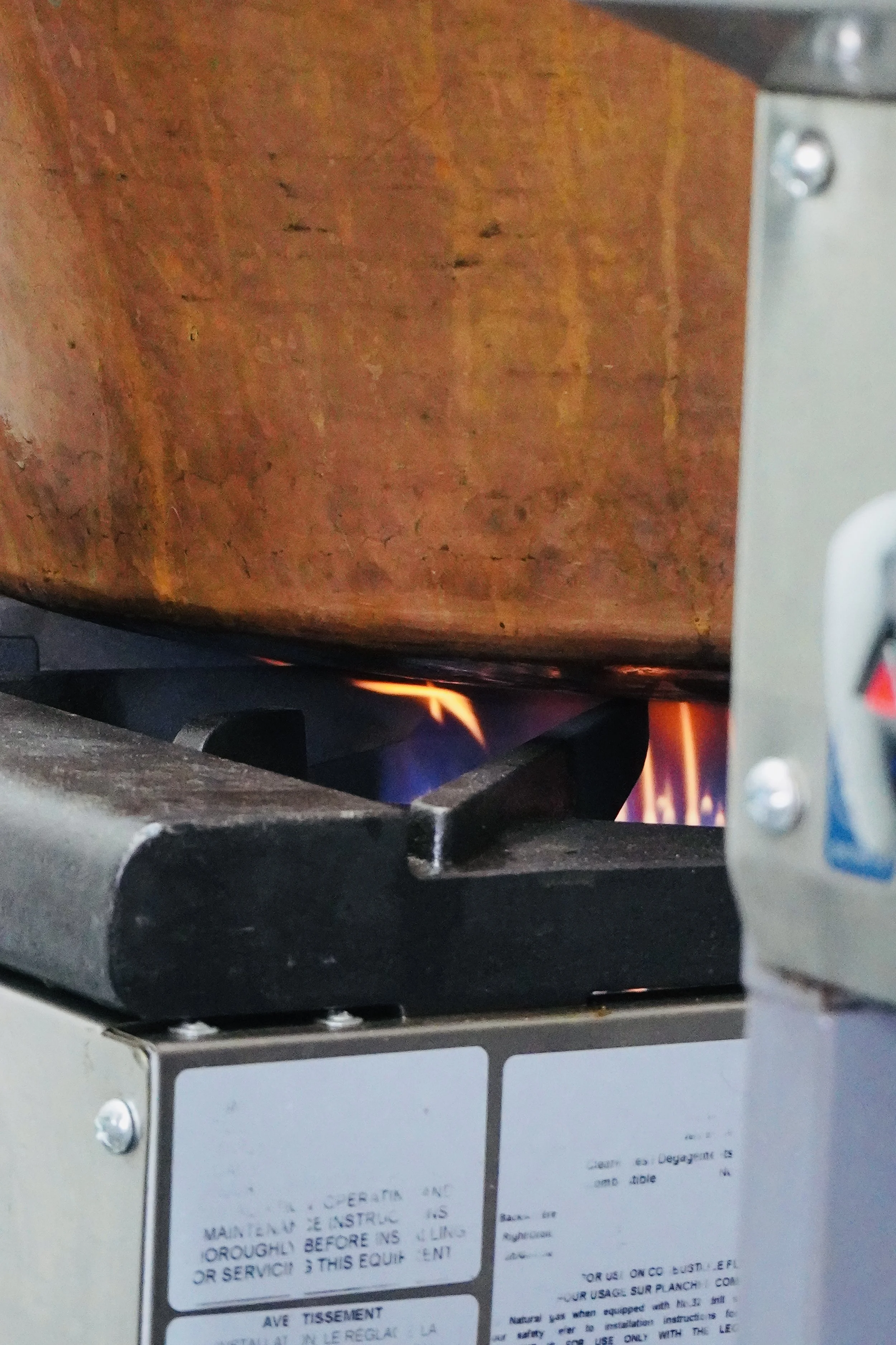 A gas stove burner with a pan on top, heating with visible flames, and a partially visible control panel and label in the foreground.