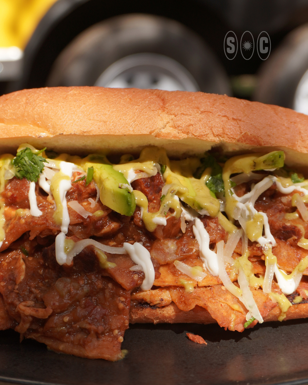 Close-up of a chilaquiles torta with shredded pork, avocado slices, shredded cheese, and cream on a toasted bun.