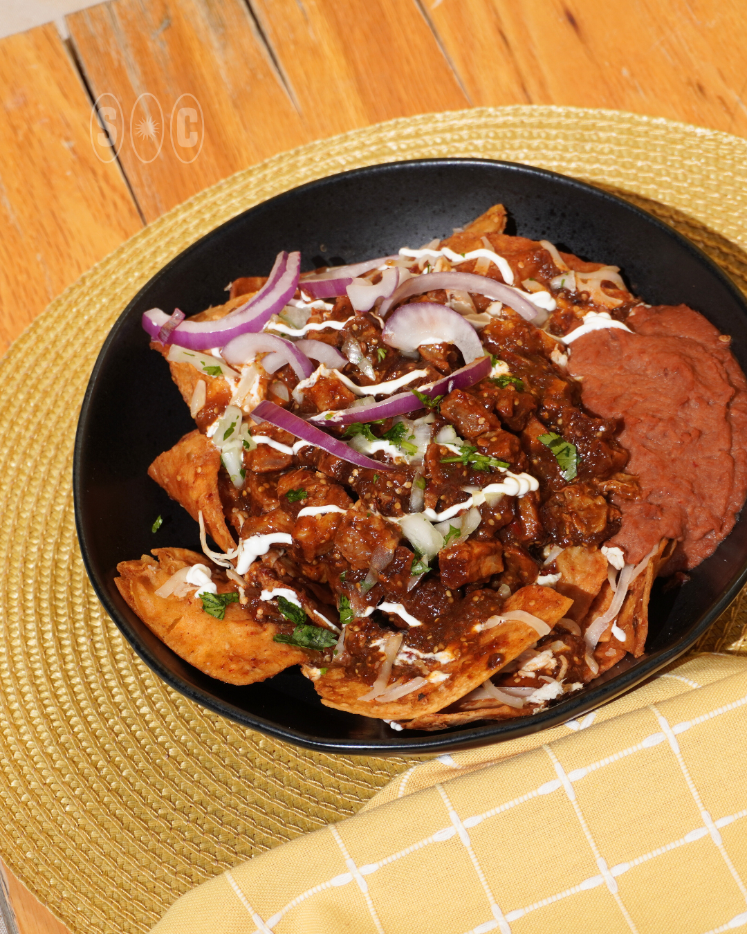 A black bowl of beef and chips with toppings including sour cream, chopped cilantro, shredded cheese, diced onions, and red sauce, placed on a woven placemat on a wooden table with a yellow cloth.