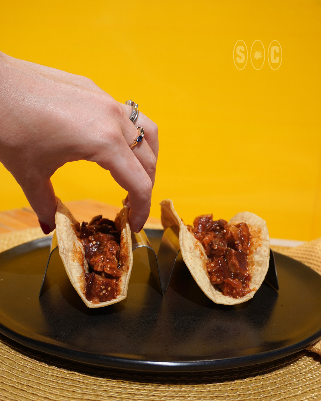 A hand with rings holds a taco filled with chicharron morita sauce, placed on a black plate against a yellow background.