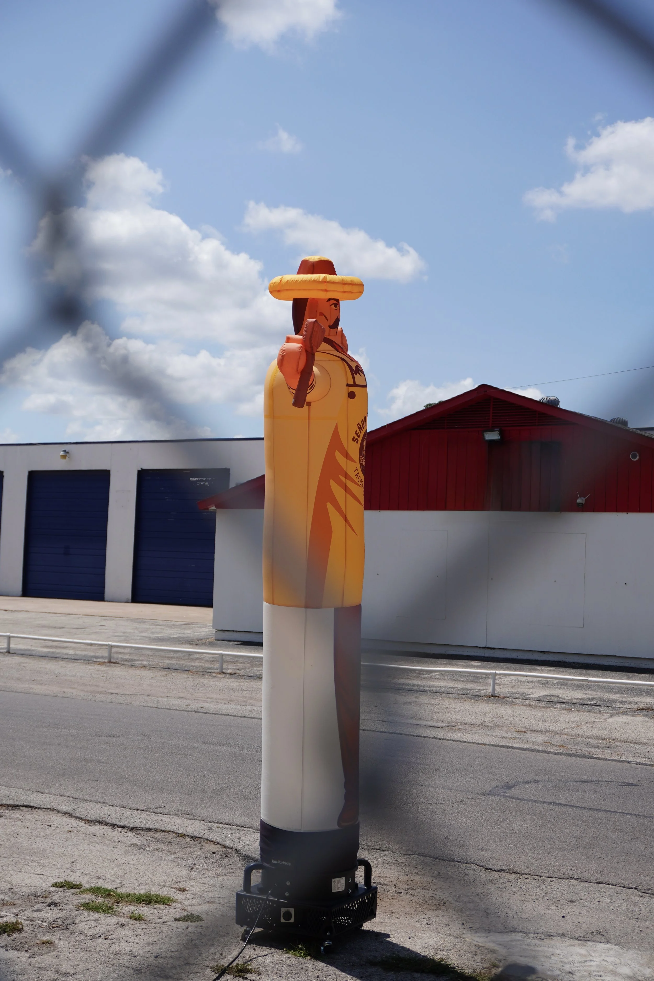 A large inflatable man wearing a sombrero and holding a wood cooking utensil, standing outdoors near a white building with blue garage doors and a red building, under a partly cloudy sky.