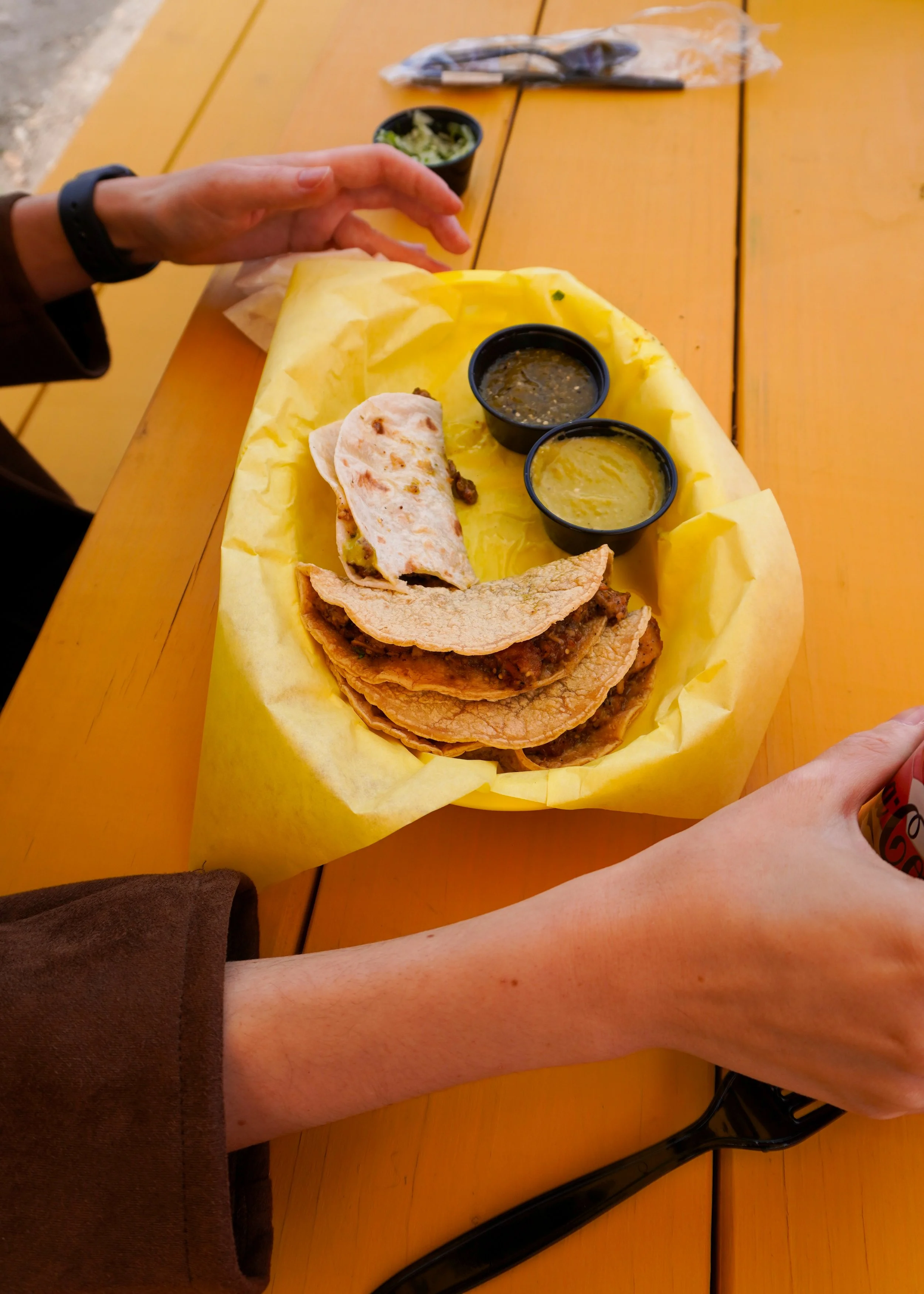 A person sitting at a yellow table enjoying Mexican tacos with two cups of salsa, with a hand reaching for a can of soda.