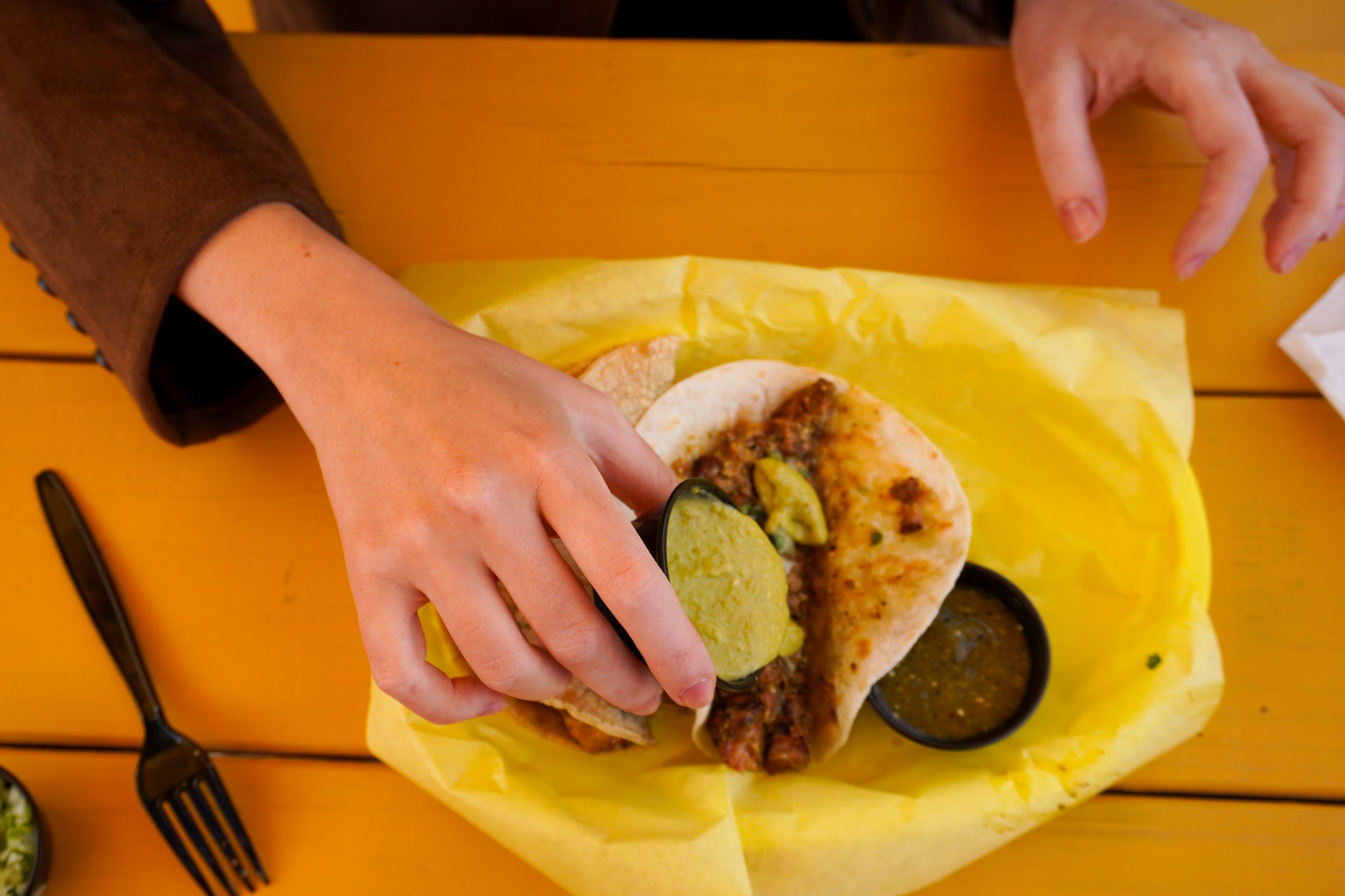 A person preparing a taco with grilled meat, sliced pickles, and green sauce, served with two small containers of sauces on a yellow paper lining on a wooden table.