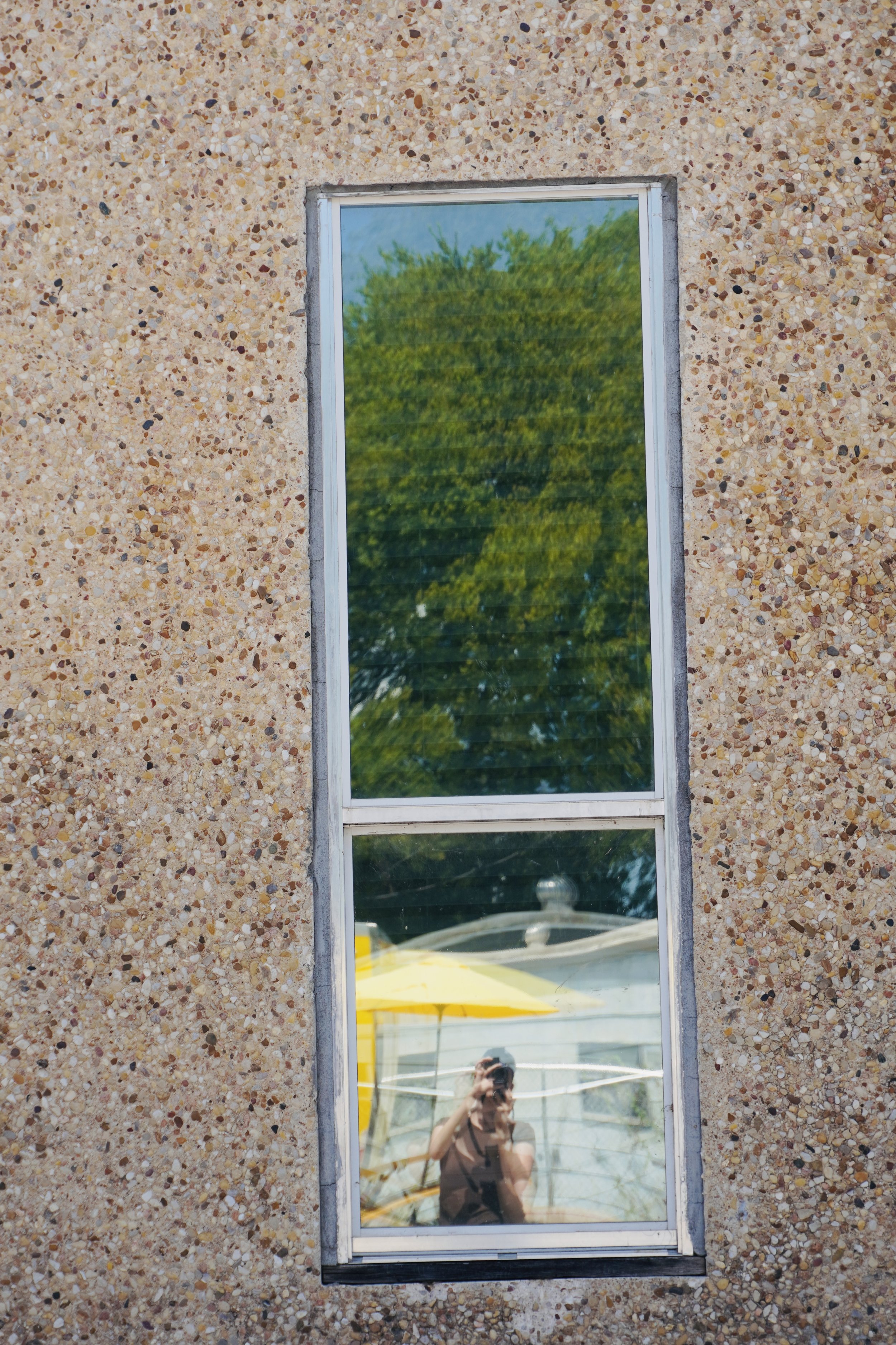 Reflection of a person taking a photo in a narrow window on a pebble-textured wall with trees and outdoor seating visible outside.