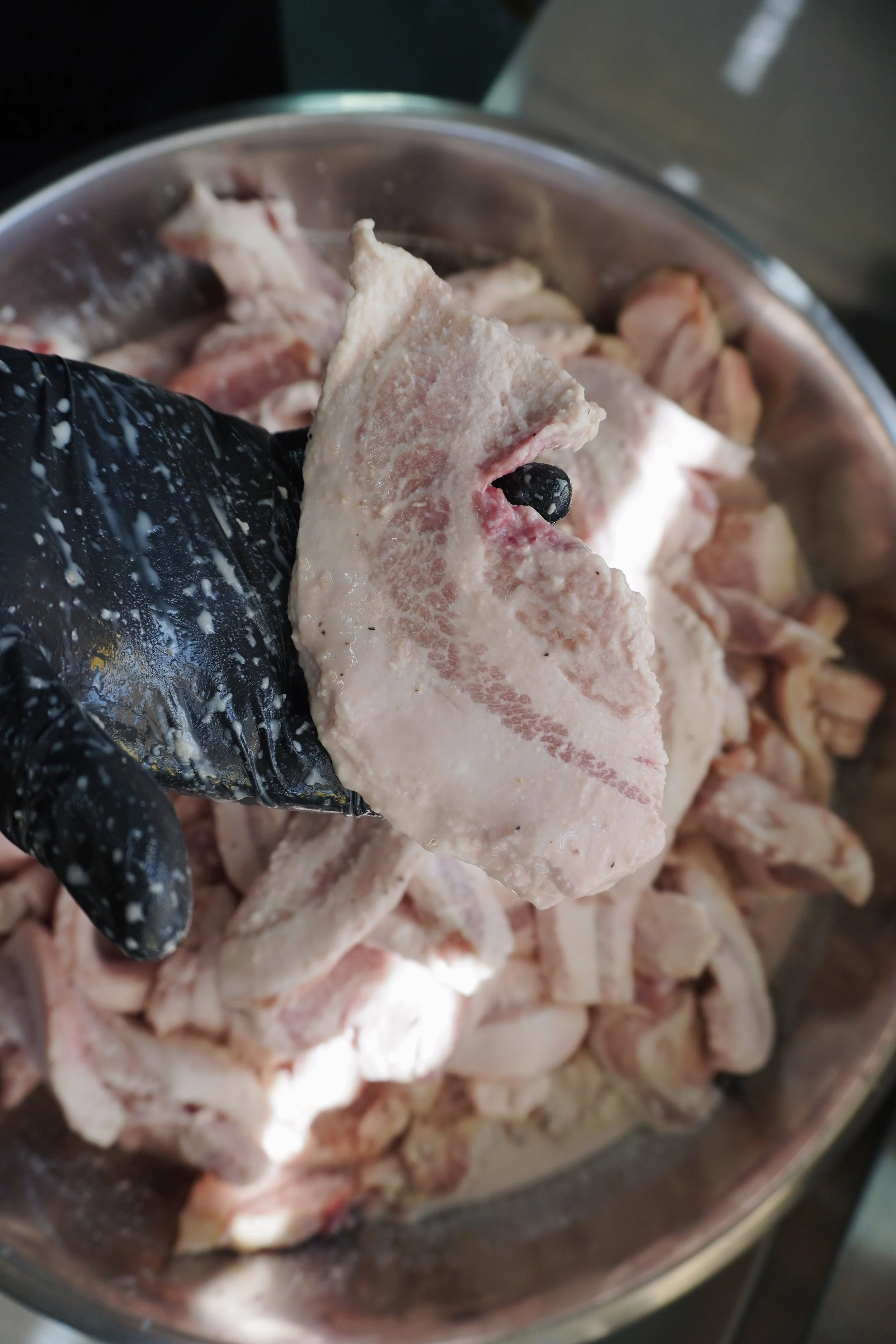 Close-up of a piece of raw pork with a black peppercorn on it, held over a bowl of raw pork pieces.