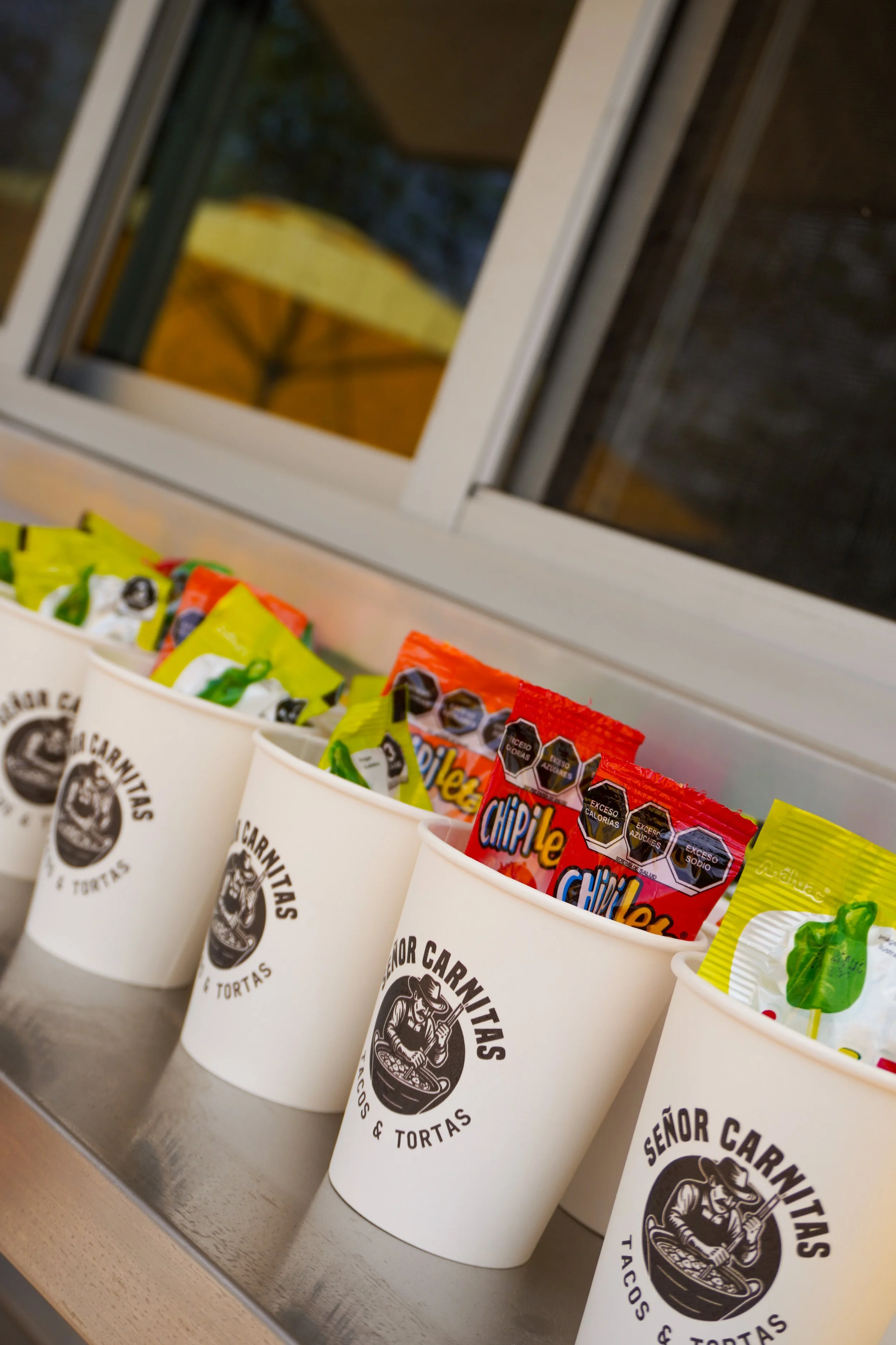 Various snack bags in white cups with a logo for Señor Carnitas, arranged on a counter with a window in the background.