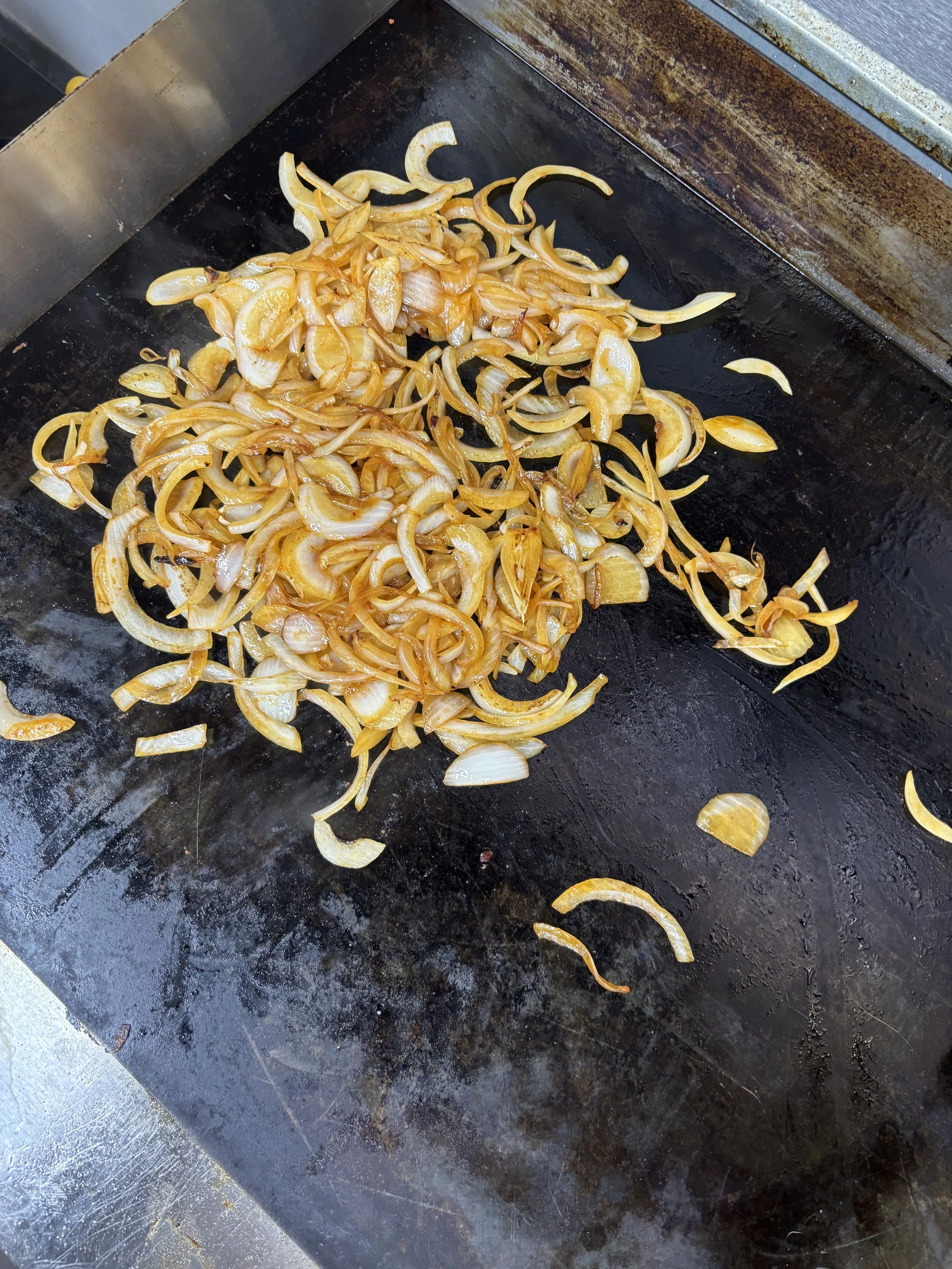 On a black griddle, sliced onions are being cooked, some caramelized to a golden brown color.