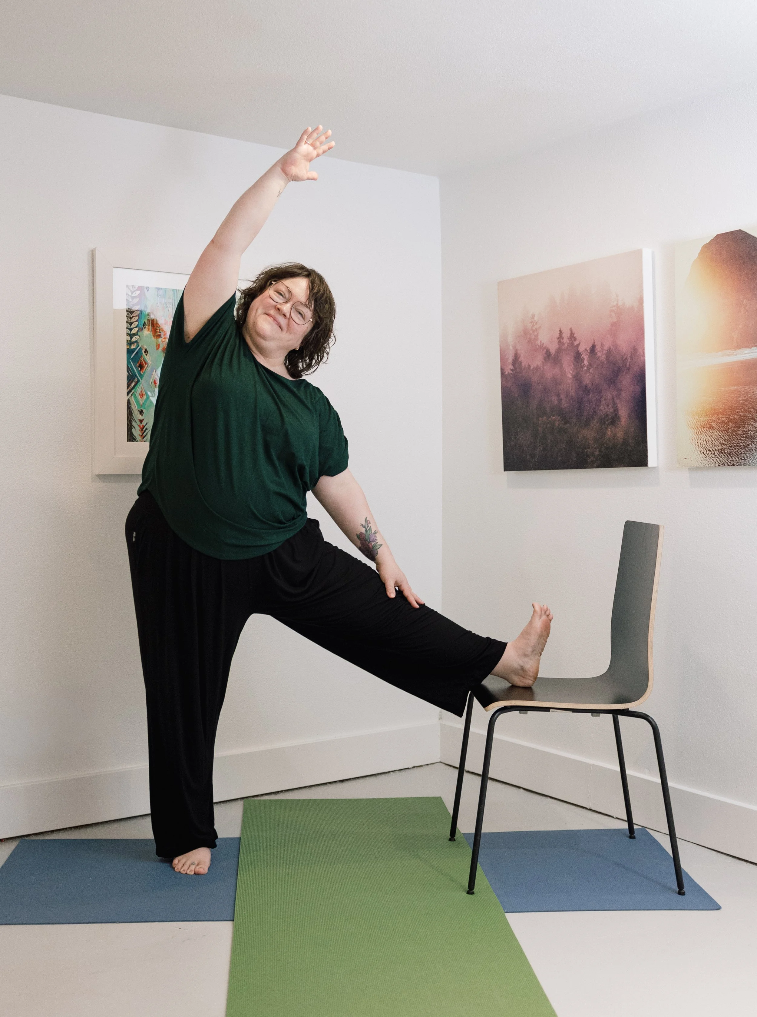 A person with short hair, glasses, and a black outfit standing on a yoga mat, stretching with one leg on a chair and one arm raised above her head in a yoga pose, in a room with nature-themed artwork on the wall.