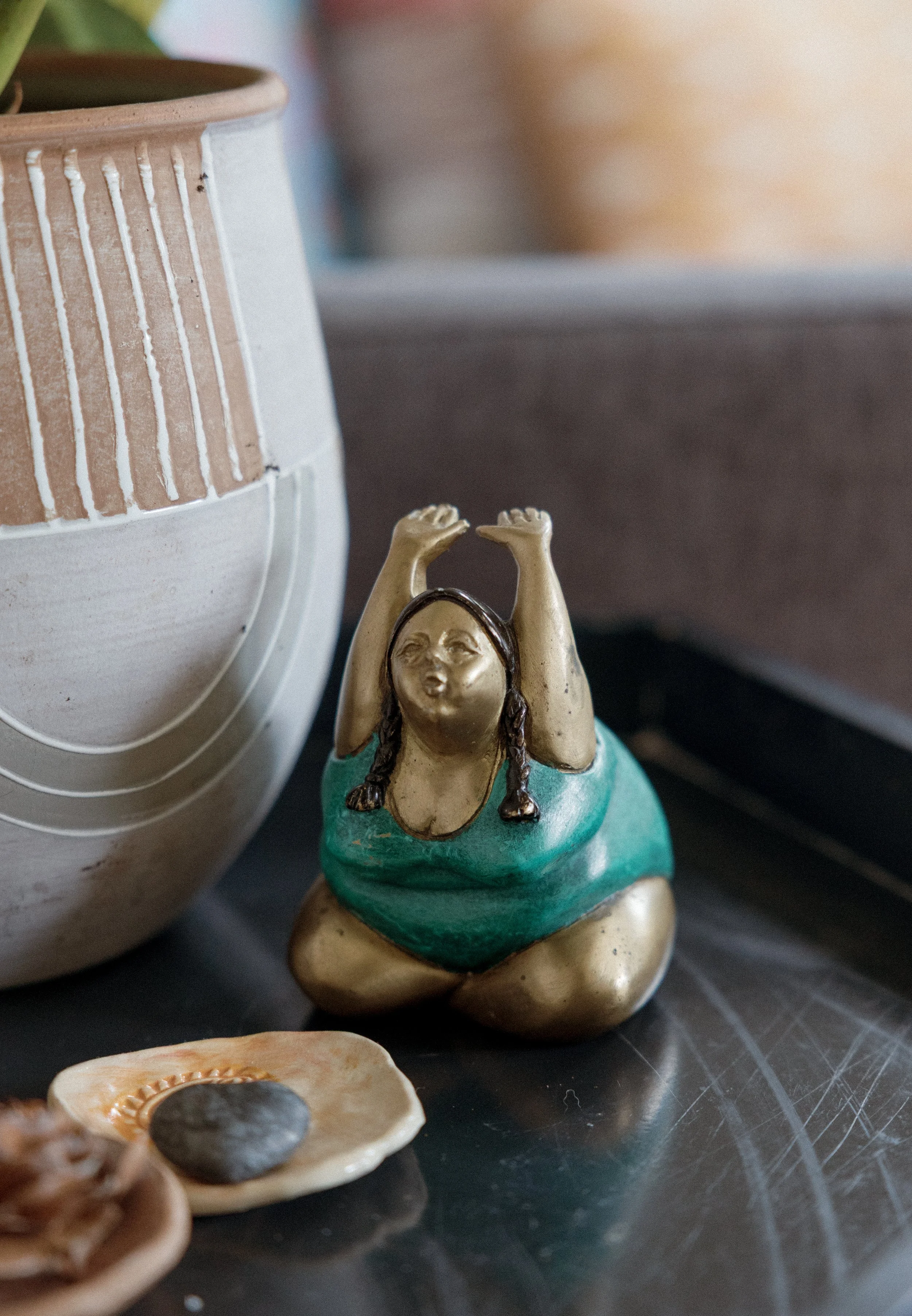 Close-up of a small figurine of a woman with braids, sitting with her hands raised above her head, wearing a turquoise dress, on a reflective surface surrounded by pottery and shells.