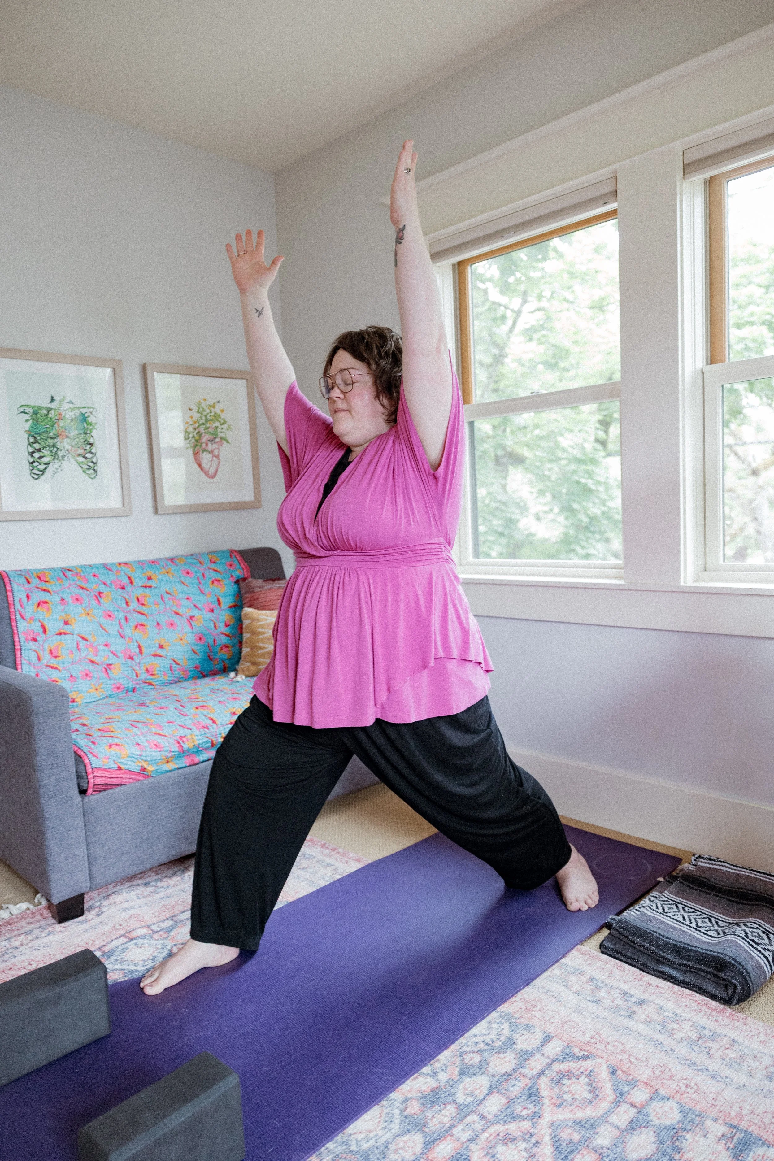 Woman practicing yoga indoors, standing in a warrior pose with arms raised, on a purple yoga mat in a room with large windows and wall art.