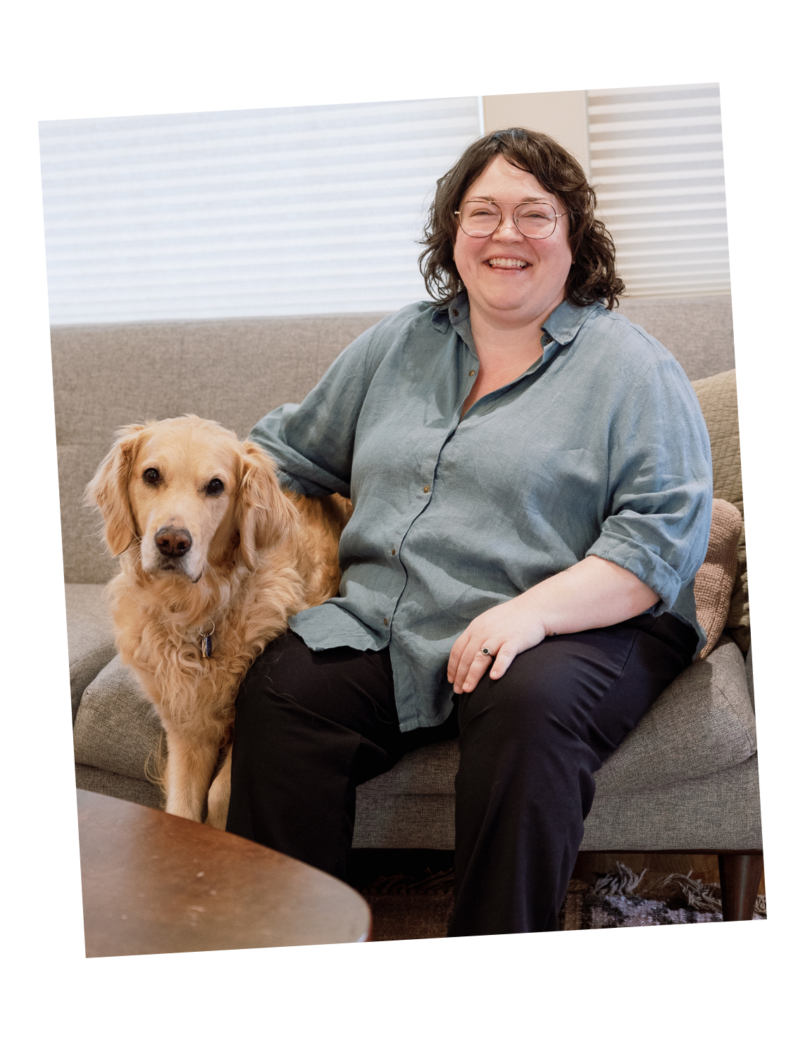 A woman with glasses sitting on a sofa, smiling, with a golden retriever dog sitting beside her.