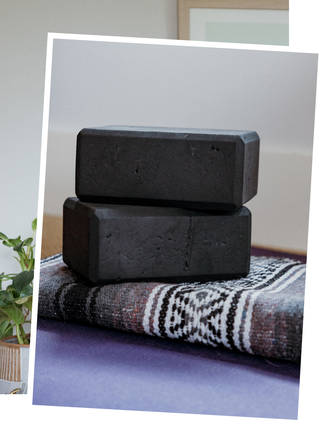 Two black yoga blocks stacked on a patterned blanket on a purple yoga mat, with a plant nearby.