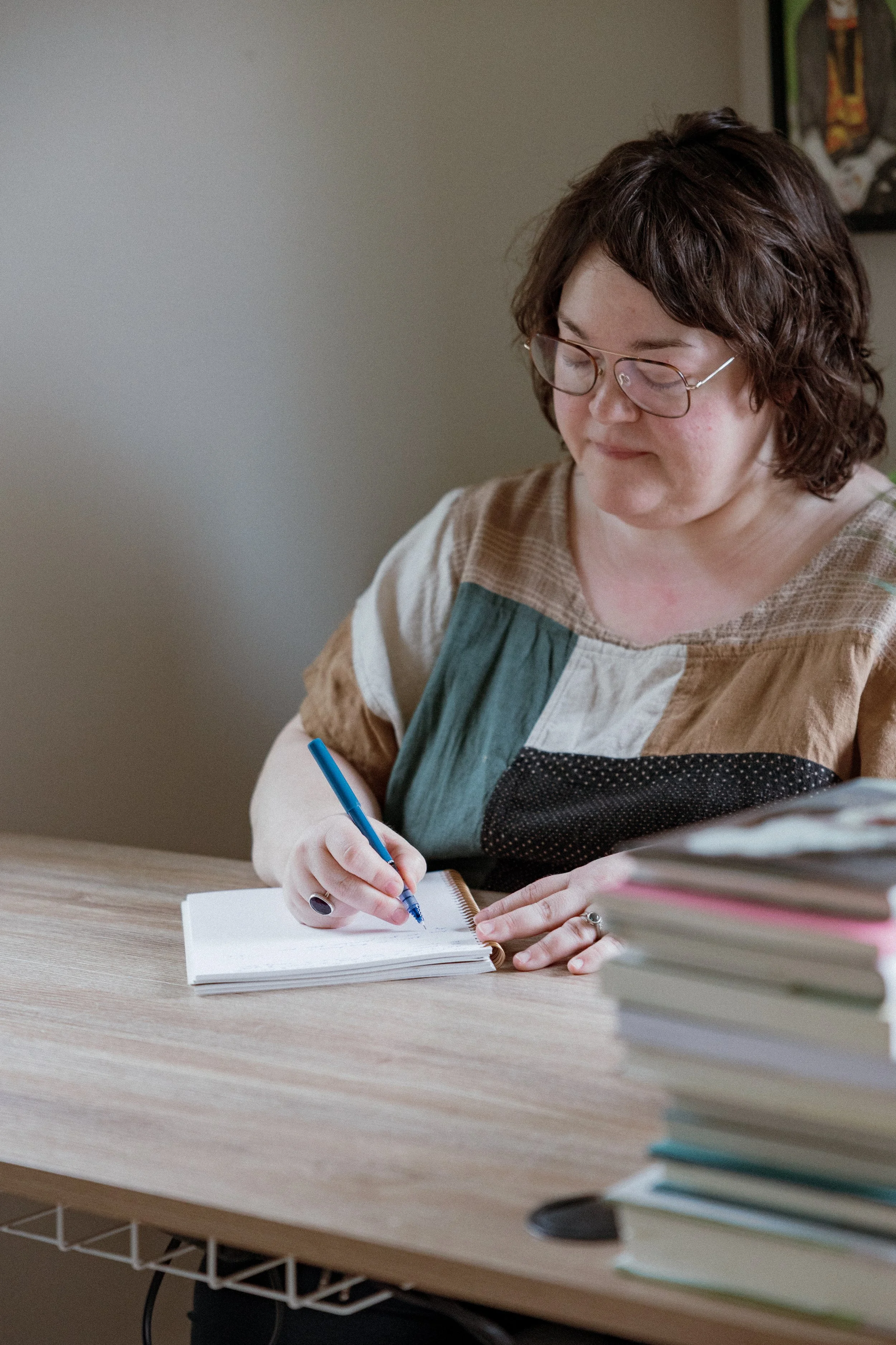 A woman with glasses and short curly brown hair is sitting at a wooden desk, writing in a small notepad with a blue pen. There are stacks of books on the right side of the desk.